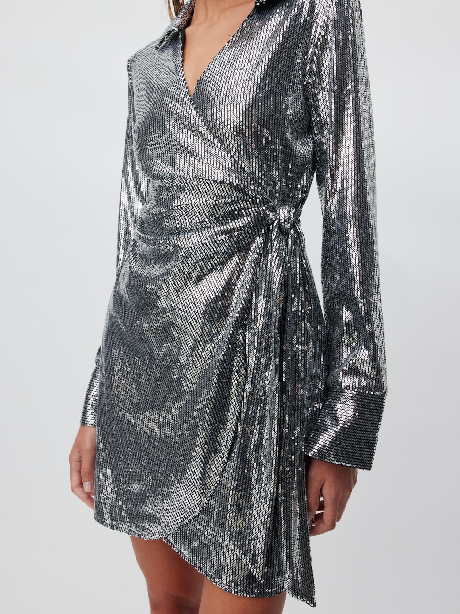 Eine Person trägt das Kleid „Meline“ von LeGer by Lena Gercke, ein schimmerndes silbernes Wickelkleid mit langen Ärmeln und einem Knotendetail an der Taille. Das Kleid hat eine metallische, plissierte Textur und eine normale Passform. Das Foto zeigt das Outfit vom Hals bis zur Mitte des Oberschenkels und zeigt die reflektierende Qualität des Kleidungsstücks.