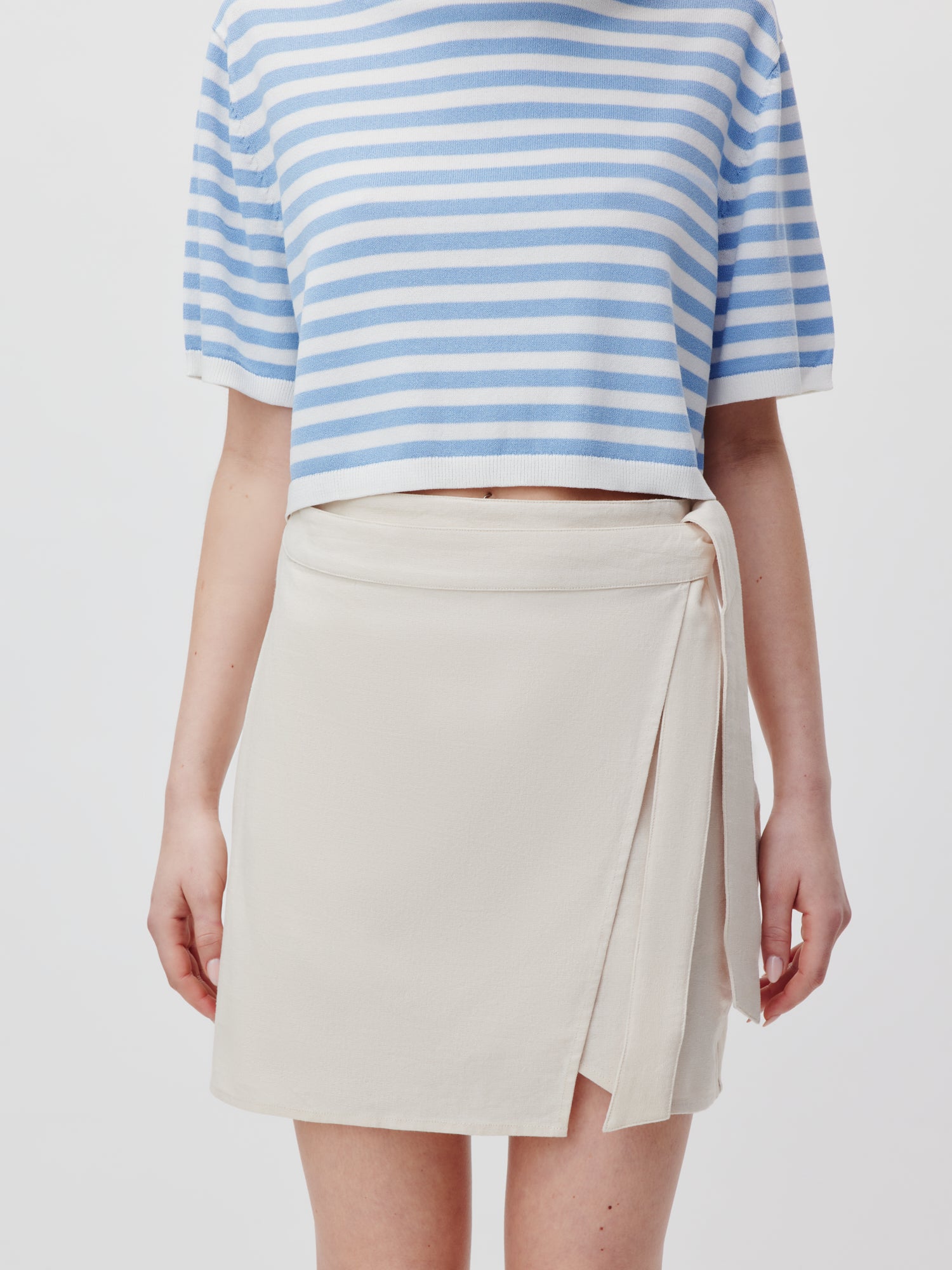 Eine Person trägt ein blau-weiß gestreiftes, kurzärmeliges Crop-Top, das mit dem Marlene Rock von LeGer by Lena Gercke in Hellbeige kombiniert wird und eine schmeichelhafte Silhouette schafft. Das Bild ist vor einem schlichten weißen Hintergrund und zeigt nicht den Kopf der Person.