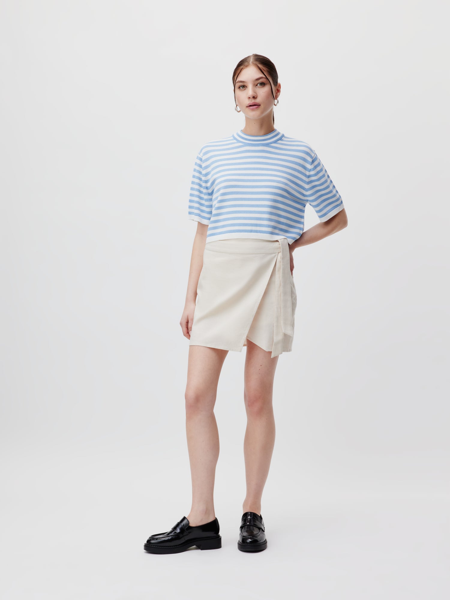Eine Frau steht vor einem schlichten weißen Hintergrund und trägt den eleganten cremefarbenen Marlene Rock von LeGer by Lena Gercke, gepaart mit einem hellblau-weiß gestreiften, kurzärmeligen Top und schwarzen Slippern. Ihr Haar ist zurückgesteckt, eine Hand ruht auf ihrer Hüfte, um eine schmeichelhafte Silhouette zu schaffen.