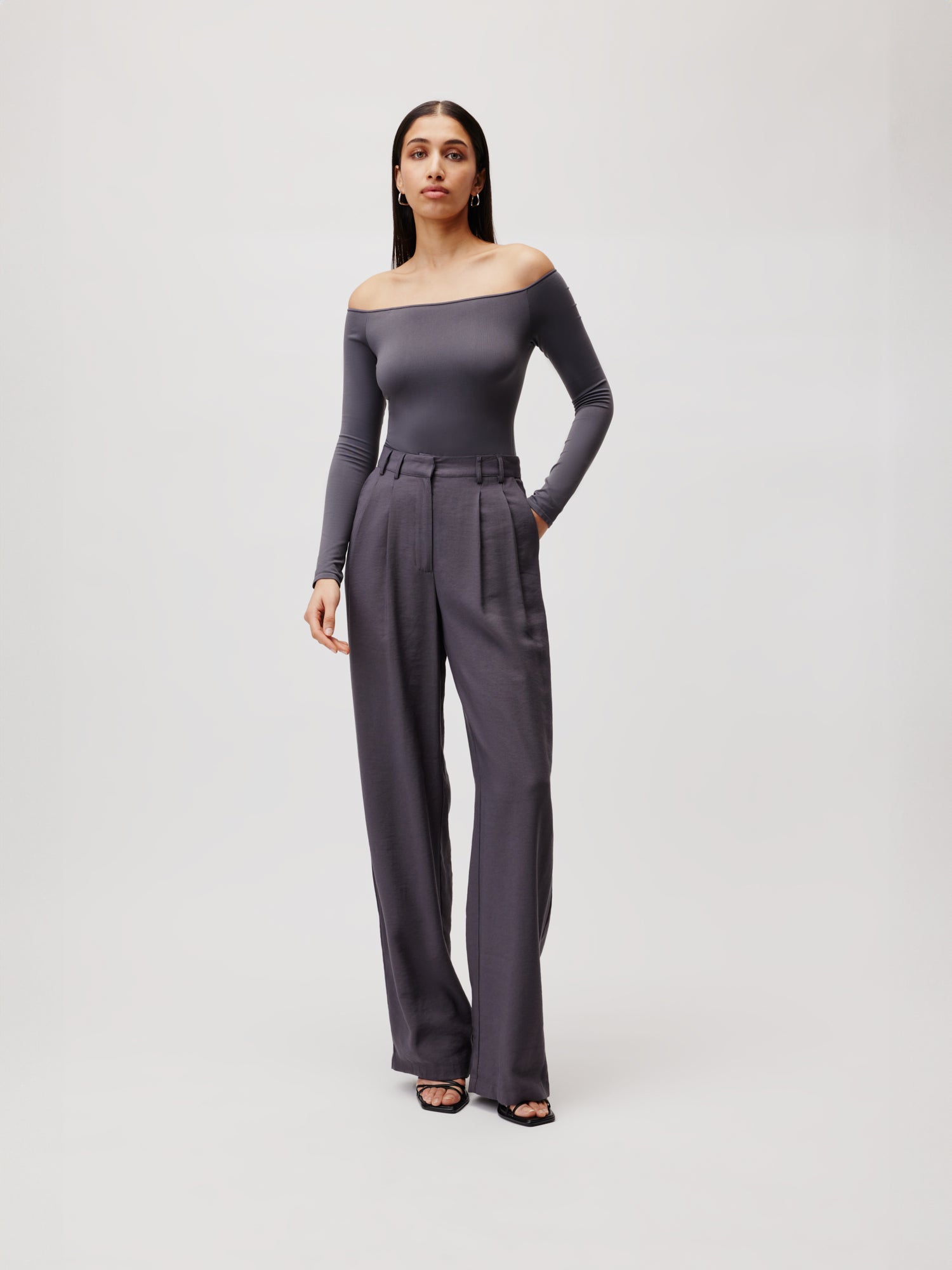 Eine Frau steht vor einem neutralen Hintergrund und präsentiert den Body Athina von LeGer by Lena Gercke. Sie kombiniert ihr langärmliges, schulterfreies graues Oberteil mit hoch taillierten, weiten dunkelgrauen Hosen. Der Look wird durch schwarze Sandalen vervollständigt. Für einen stilvollen Auftritt lässt sie ihr Haar natürlich fallen.