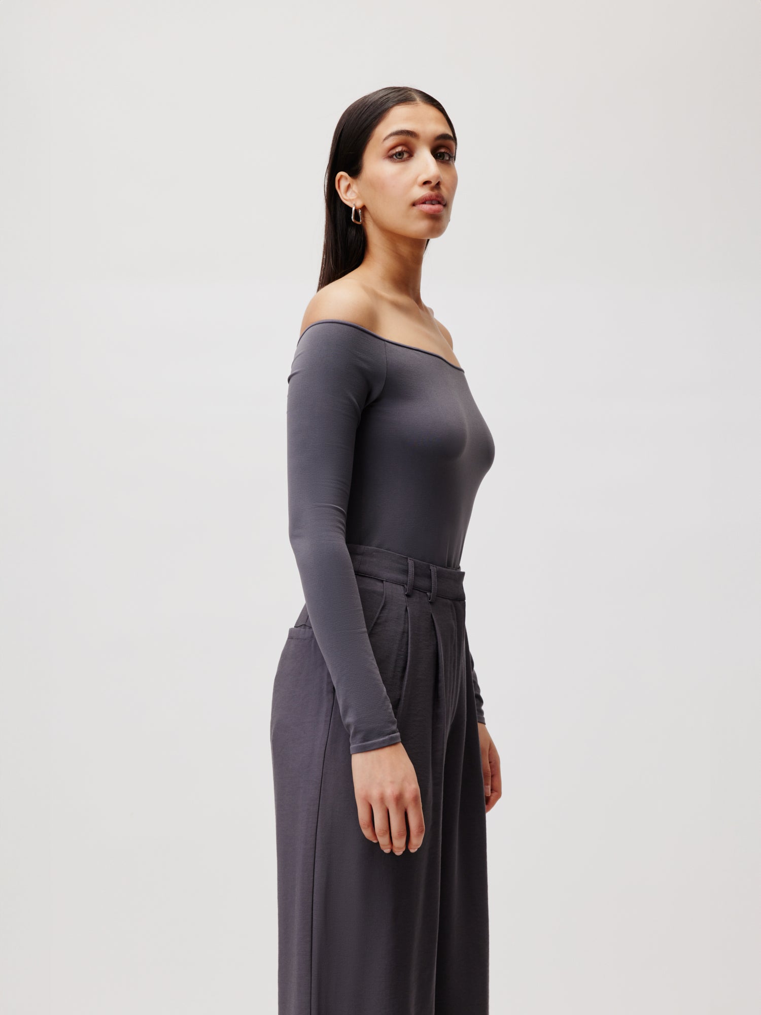 Eine Person mit langen dunklen Haaren steht im Profil vor einem schlichten Hintergrund. Sie trägt den Body Athina, ein tailliertes, schulterfreies graues Oberteil von LeGer by Lena Gercke, kombiniert mit einer hoch taillierten grauen Hose. Der Ausdruck der Person ist neutral und vermittelt Ruhe und zeitlose Eleganz.