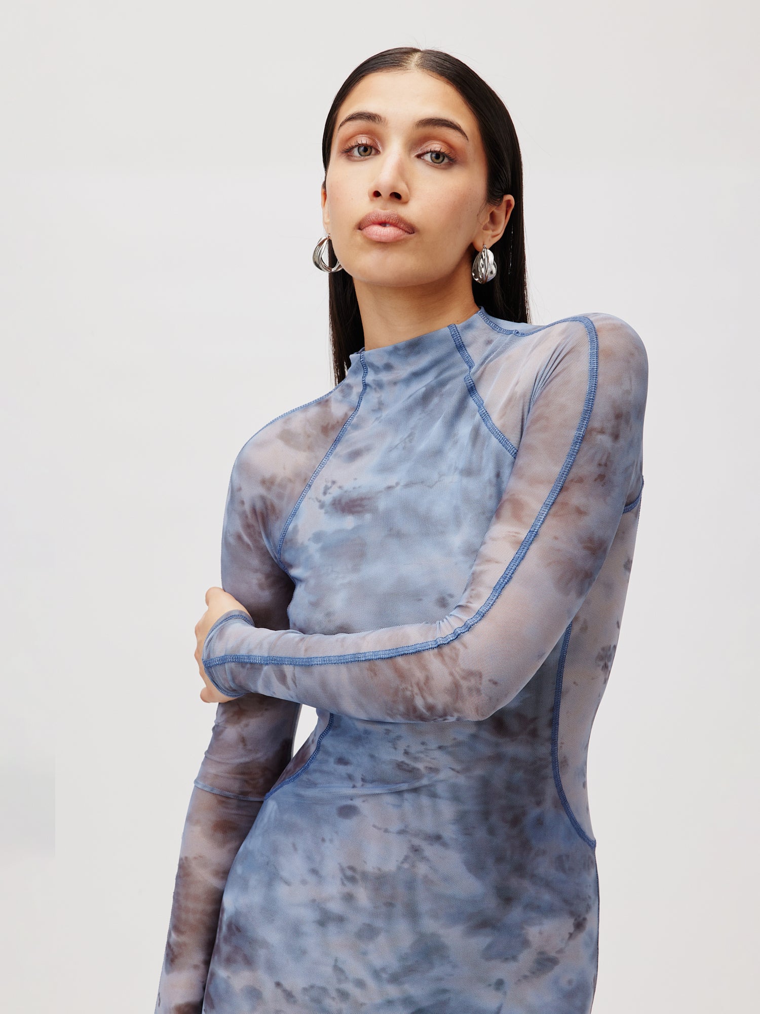 Eine Person mit langen dunklen Haaren steht selbstbewusst da und trägt das Kleid Daliah von LeGer by Lena Gercke, ein 3/4-langes blaugraues Batikkleid mit transparenten Einsätzen und langen Ärmeln. Sie ist mit Creolen geschmückt und blickt vor einem hellen Hintergrund direkt in die Kamera.
