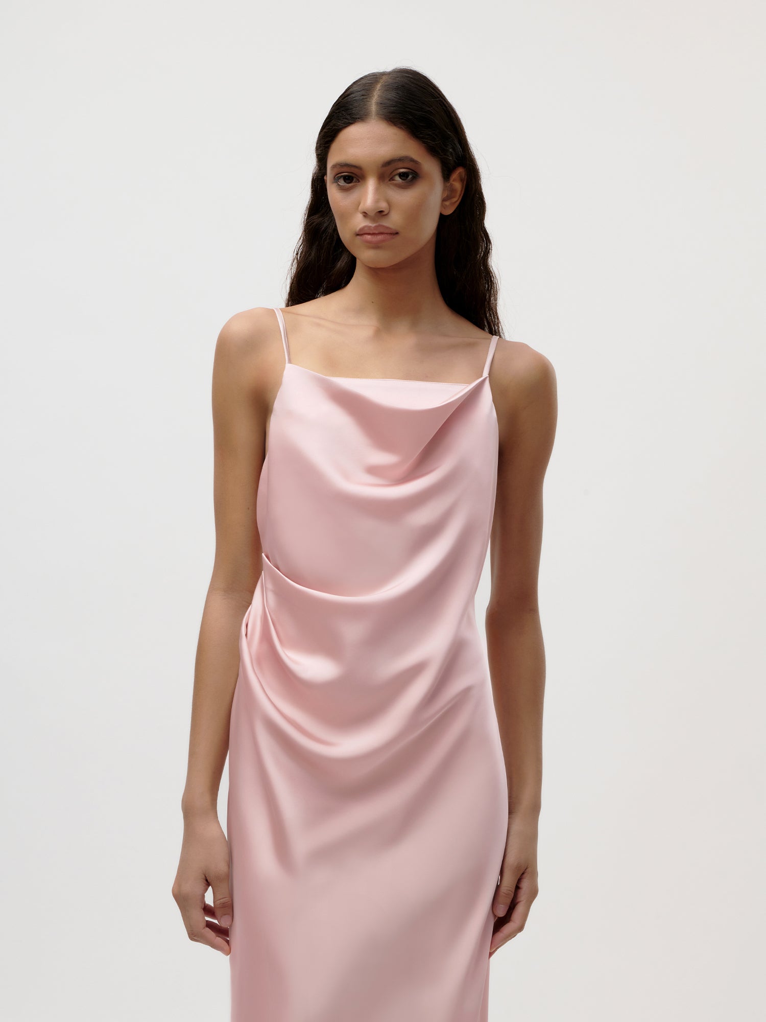 Eine Frau mit langen, gewellten braunen Haaren trägt das Brianne Dress von LeGer by Lena Gercke - ein ärmelloses, hellrosa Maxikleid aus Satin mit dünnen Trägern und einem drapierten Ausschnitt - vor einem schlichten weißen Hintergrund.