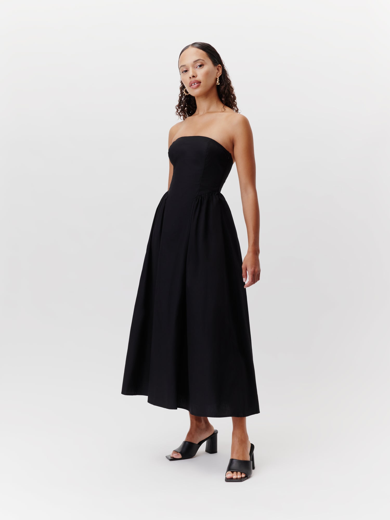 Eine Person in einem trägerlosen, schwarzen 7/8-Kleid der Marke „LeGer by Lena Gercke“ mit dem Modell „Vianne“, das ein eng anliegendes Oberteil und einen ausgestellten Rock aufweist, kombiniert mit schwarzen Sandalen mit Absatz, steht selbstbewusst vor einem schlichten weißen Hintergrund.