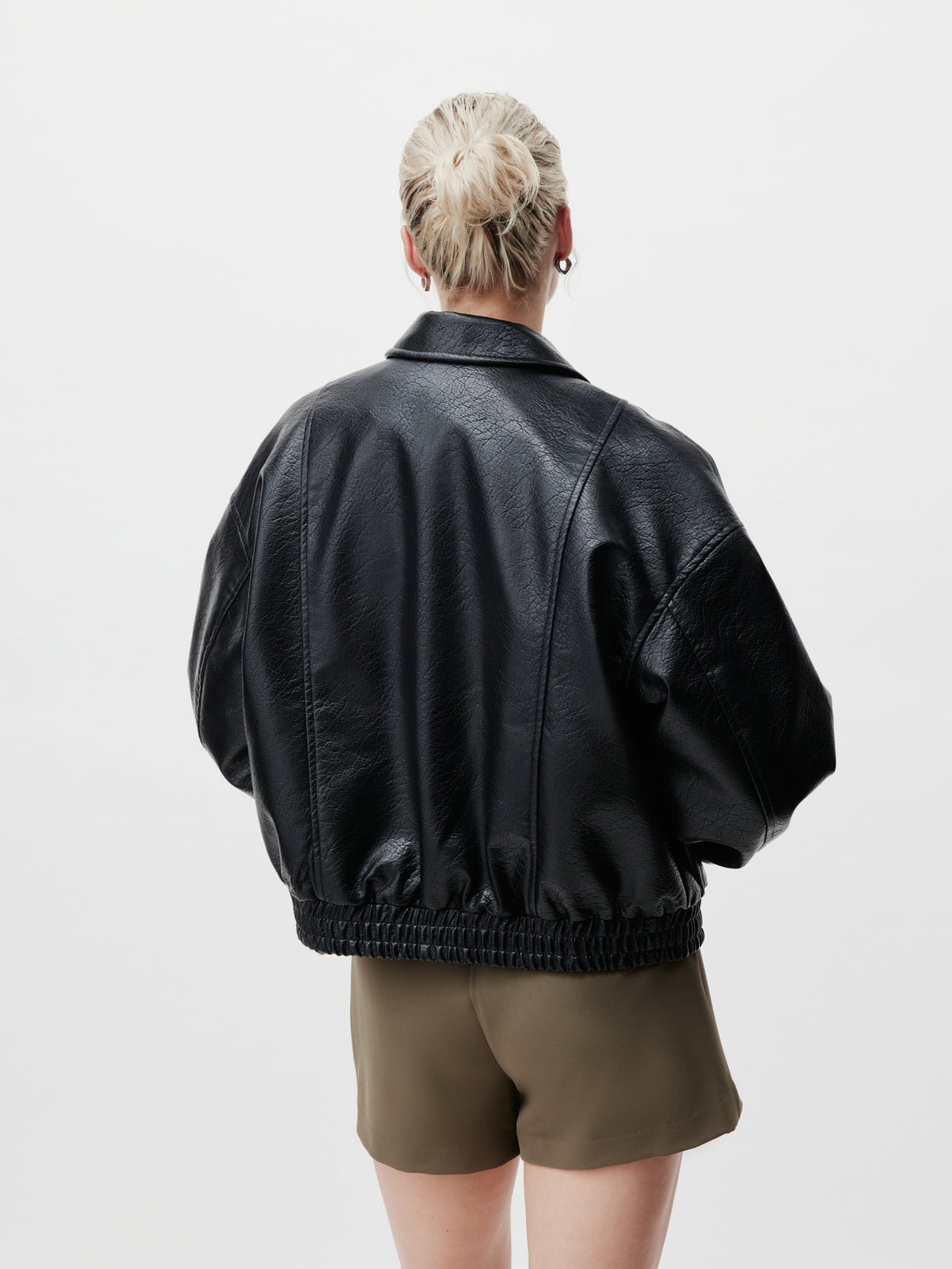 Eine Person mit blonden Haaren, die zu einem Dutt zusammengebunden sind, ist von hinten zu sehen. Sie steht vor einem schlichten weißen Hintergrund und trägt die schwarze Lederjacke „Taira“ von LeGer by Lena Gercke in Größe 36 mit langen Ärmeln, die locker über olivgrünen Shorts getragen wird.