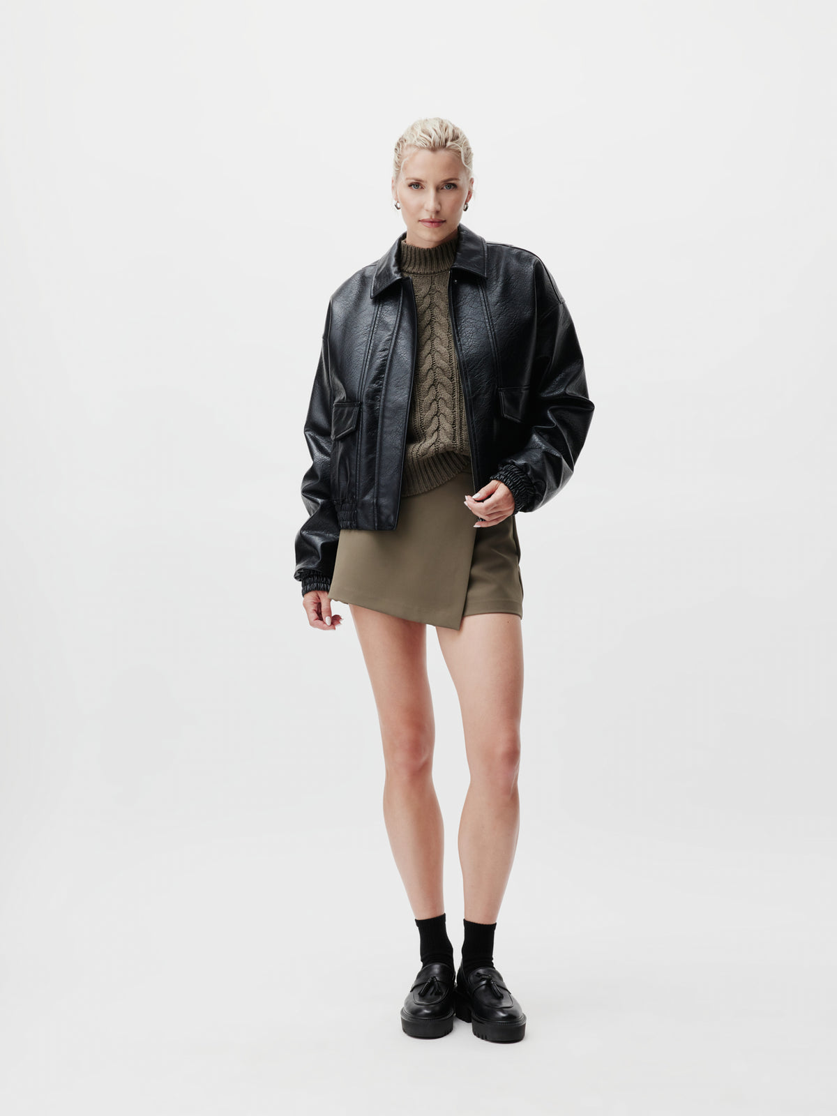 Eine Person steht vor einem weißen Hintergrund und trägt die schwarze „Taira“-Jacke von LeGer by Lena Gercke mit langen Ärmeln und normaler Länge, kombiniert mit einem khakifarbenen Strickpullover, einem khakifarbenen Minirock, schwarzen Socken und schwarzen Schuhen in Größe 36.