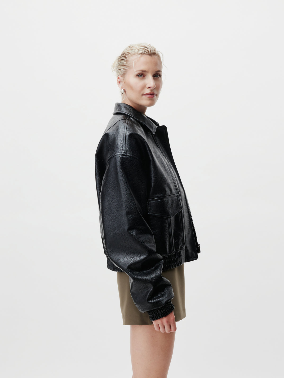 Eine Person mit blonden Haaren, die zu einem Dutt zusammengebunden sind, trägt die schwarze Lederjacke „Taira“ von LeGer by Lena Gercke in Größe 36 und einen olivgrünen Rock in normaler Länge und steht seitlich vor einem schlichten weißen Hintergrund.