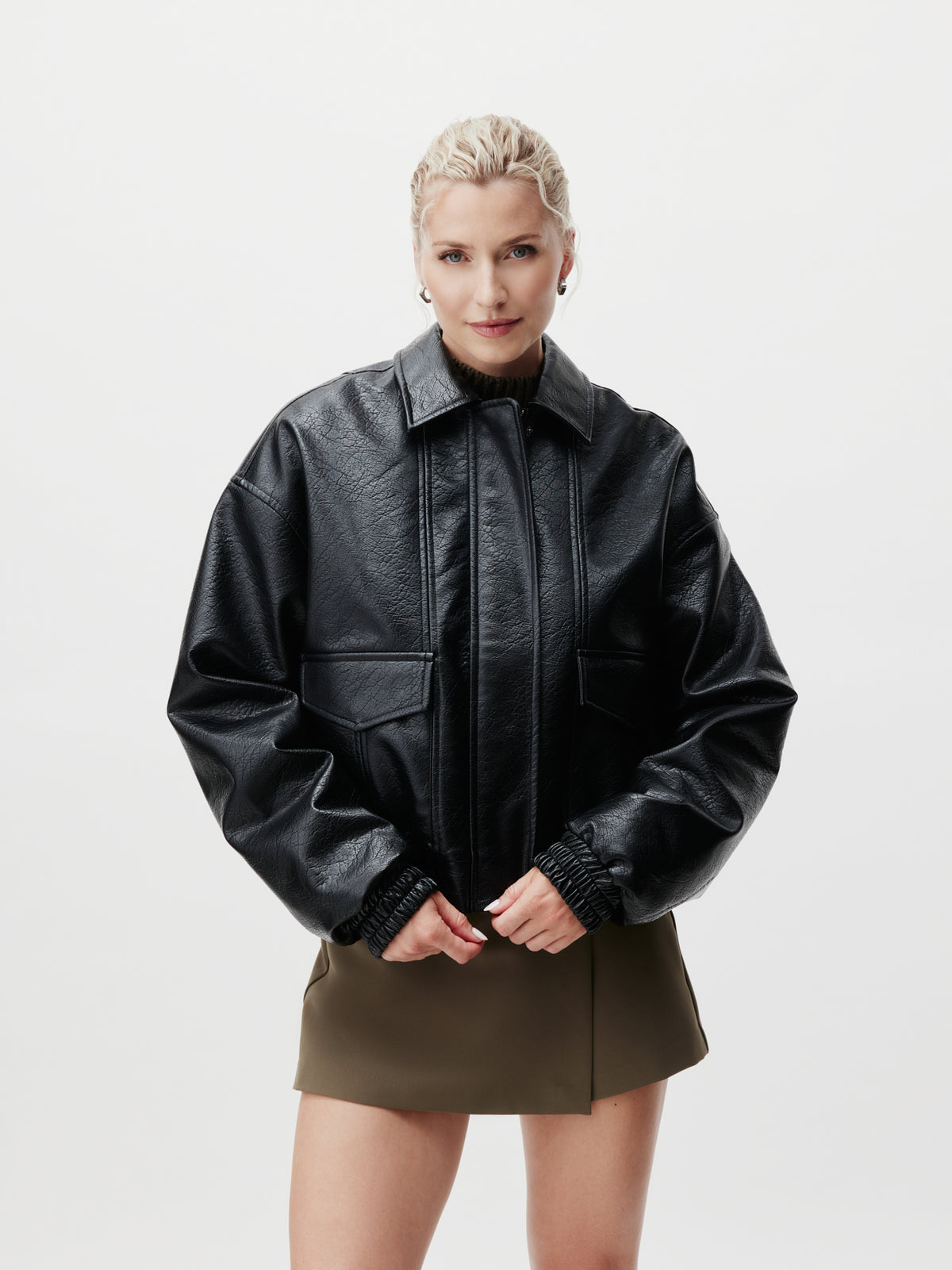 Eine Person mit blondem, zu einem Zopf geflochtenem Haar trägt die Jacke „Taira“ von LeGer by Lena Gercke – aus schwarzem Leder mit langen Ärmeln – kombiniert mit einem olivgrünen Rock in Größe 36 und steht vor einem schlichten weißen Hintergrund.