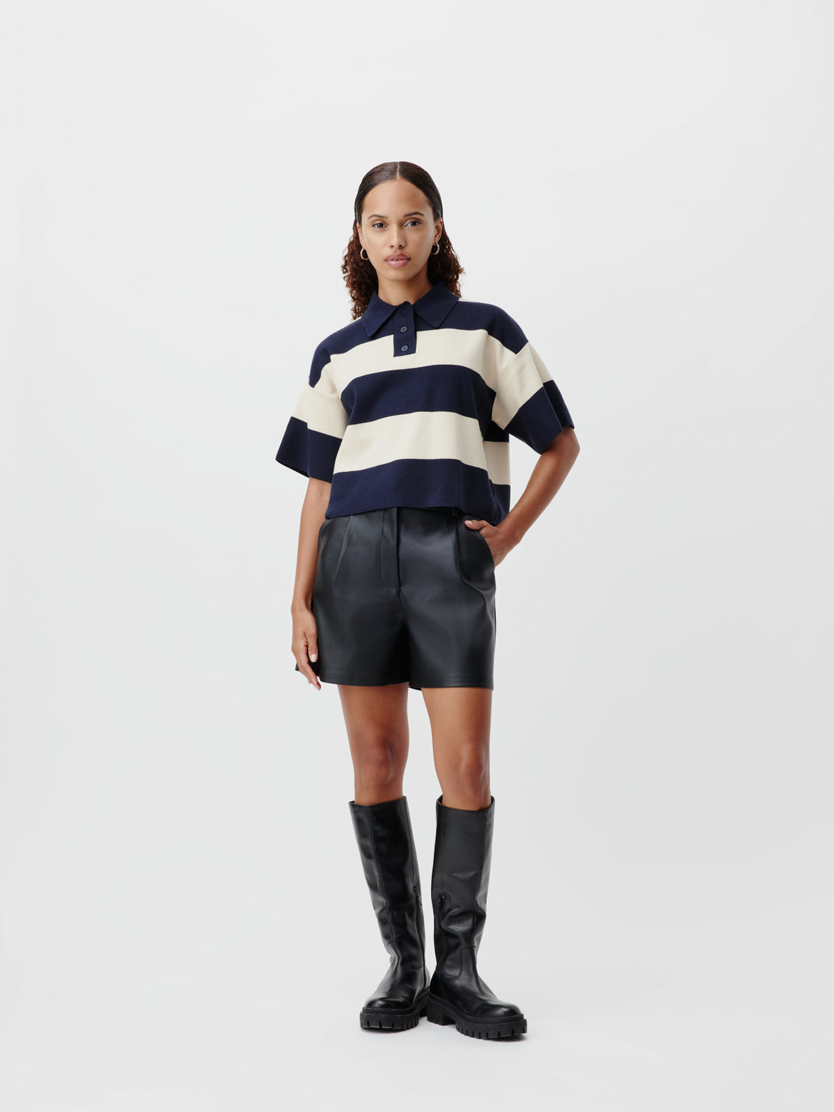 Eine Person, die ein marineblau-cremefarben gestreiftes Poloshirt, „Cay“-Shorts und schwarze kniehohe Stiefel trägt.