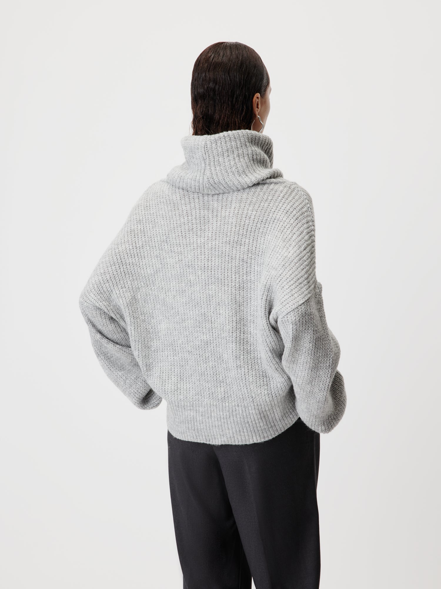 Eine Person mit dunklem, zurückgegeltem Haar ist von hinten zu sehen, die den Jarine Jumper von LeGer by Lena Gercke trägt - einen hellgrauen, gerippten Rollkragenpullover mit lockerer Passform und normaler Länge - und dazu eine schwarze Hose vor einem schlichten weißen Hintergrund.