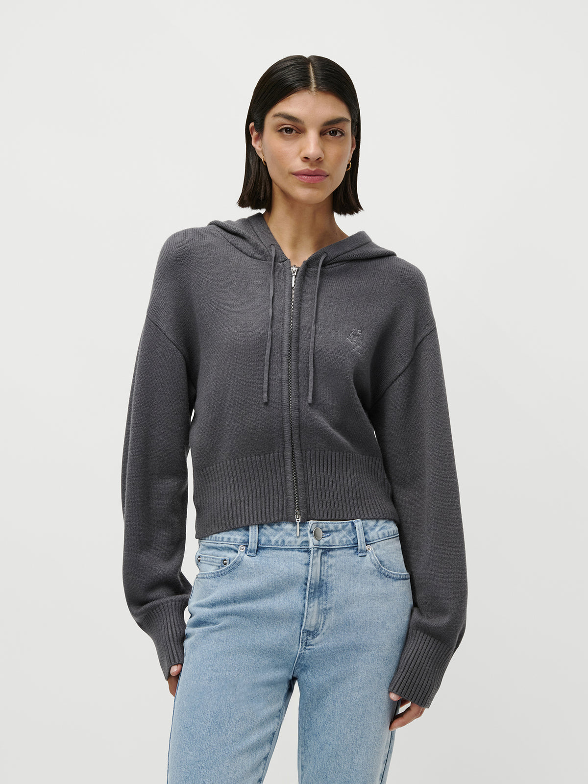 Eine Frau mit glattem, dunklem Haar trägt einen grauen LeGer by Lena Gercke „Cornelia“-Cardigan-Hoodie mit Reißverschluss und hellblaue Jeans. Sie steht vor einem schlichten weißen Hintergrund und blickt mit ausdruckslosem Gesicht in die Kamera. Dieses von Loungewear inspirierte Outfit spiegelt Lena Gercke’s lässigen Stil wider.