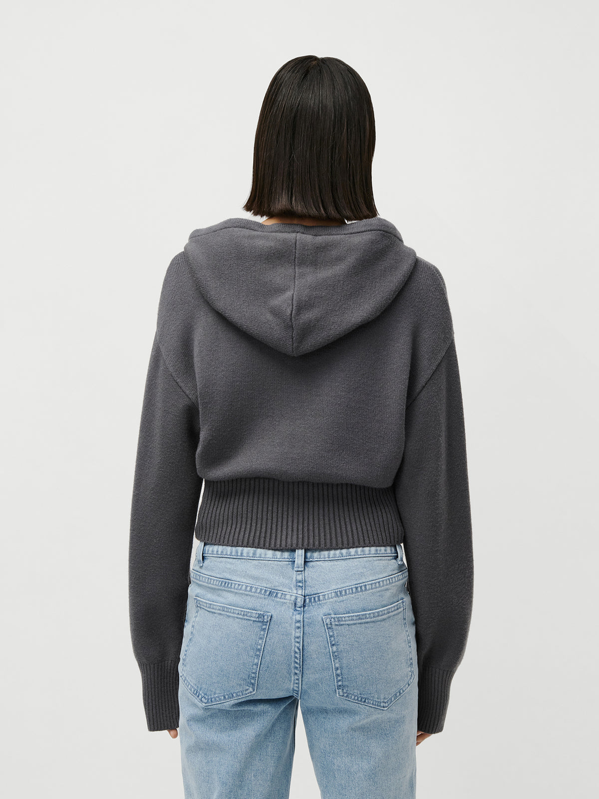 Eine Person mit glattem, dunklem Haar, gekleidet in eine dunkelgraue „Cornelia“-Strickjacke von LeGer by Lena Gercke und hellblaue Jeans, steht mit dem Rücken zur Kamera vor einem schlichten, hellen Hintergrund und verkörpert dabei mühelos die entspannte Atmosphäre moderner Loungewear.