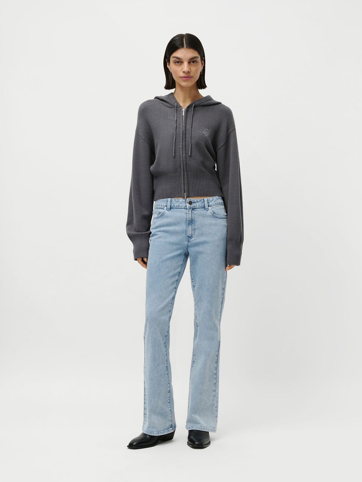 Eine Person blickt nach vorne und trägt den Cardigan „Cornelia“ aus der LeGer by Lena Gercke-Kollektion in Dunkelgrau, der als gemütliche Loungewear gestylt ist. Dazu trägt sie hellblaue Jeans und schwarze Schuhe vor einem schlichten weißen Hintergrund.