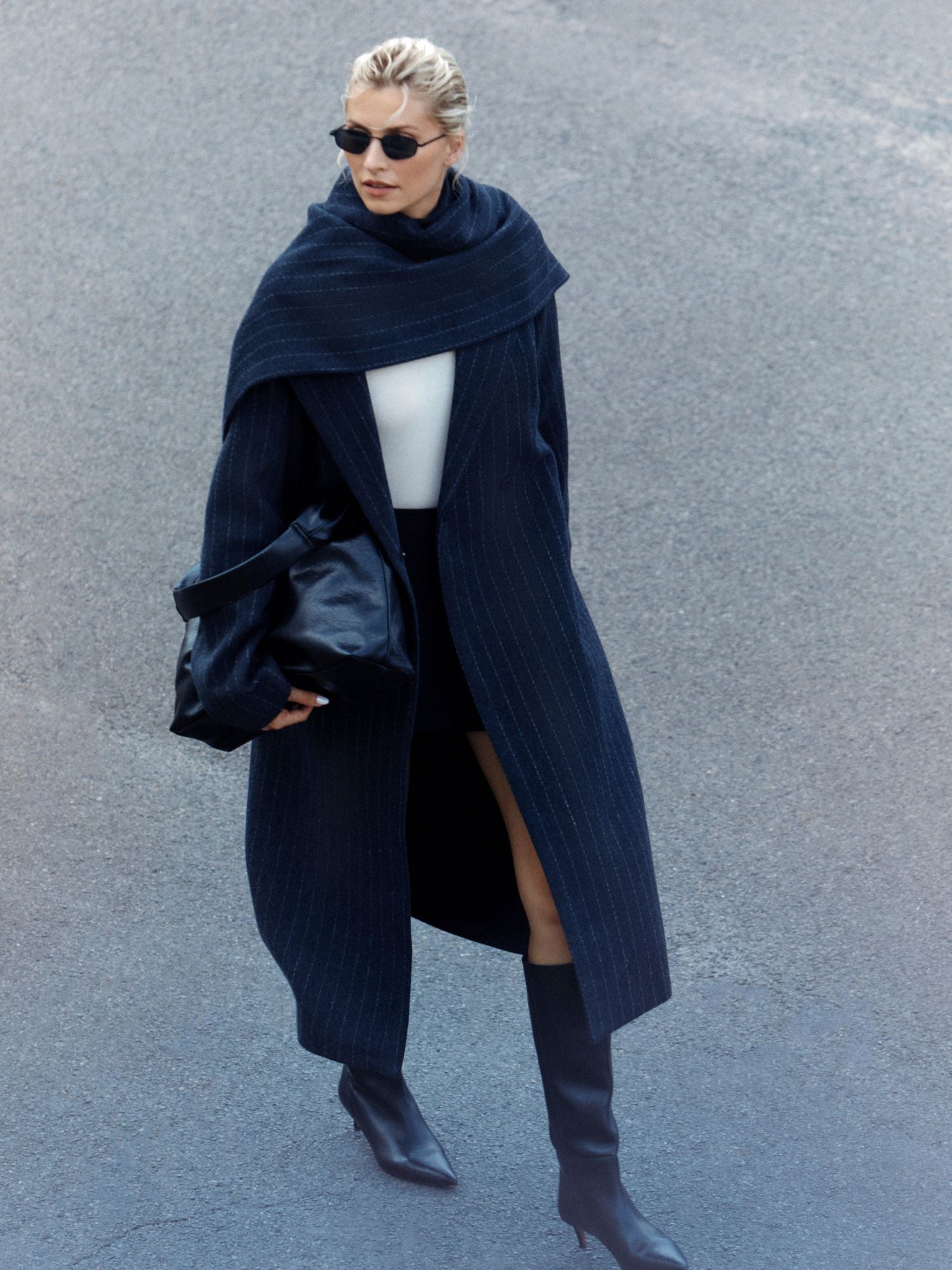 Eine Frau mit Sonnenbrille, einem LeGer by Lena Gercke Jaden Coat, schwarzem Schal, weißem Top, kurzem schwarzem Rock und kniehohen schwarzen Stiefeln geht auf dem Bürgersteig und trägt eine große schwarze Tasche.