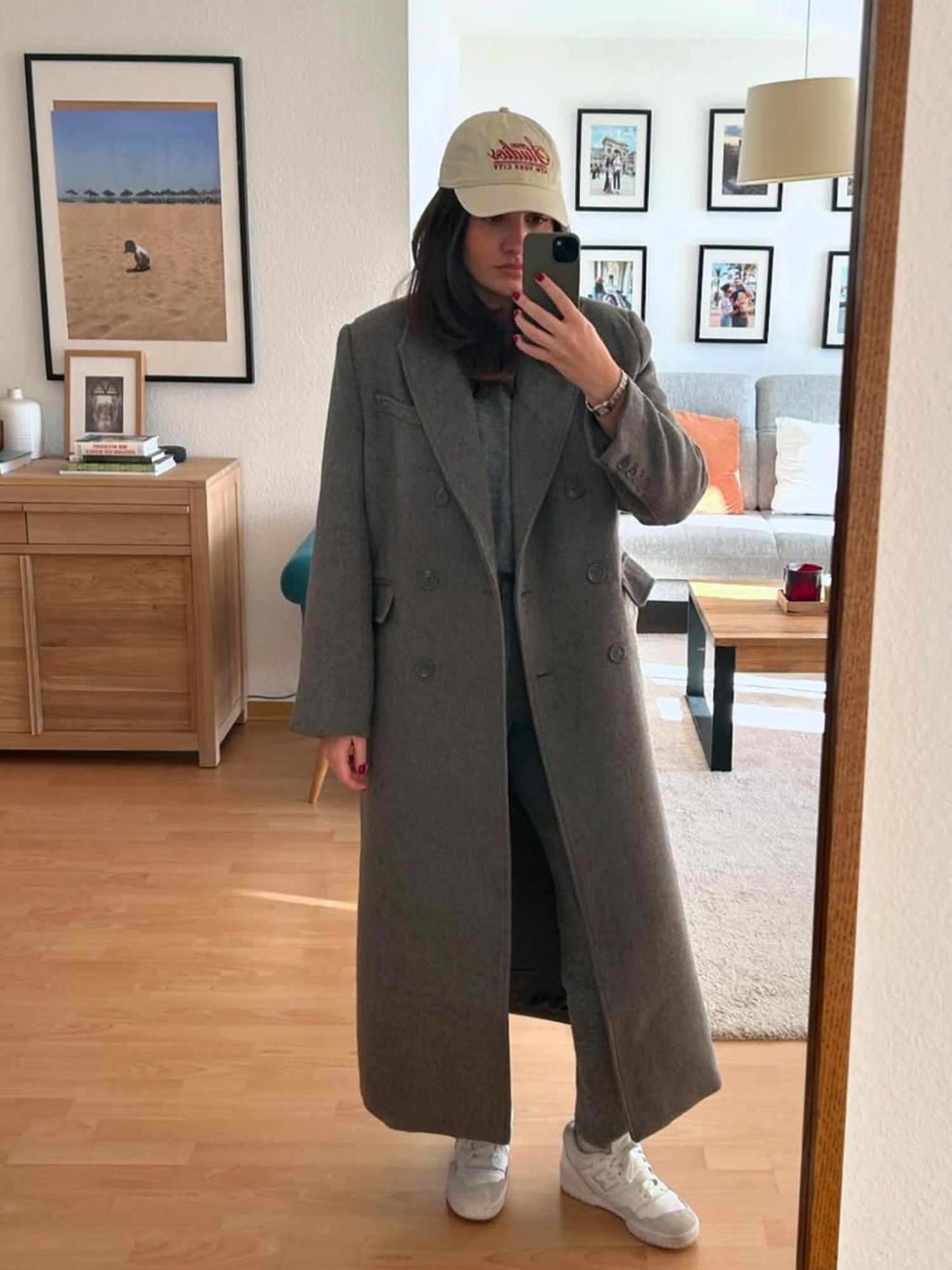 Eine Frau macht ein Spiegel-Selfie im Wohnzimmer und trägt dazu den langen grauen Kitty Coat von LeGer by Lena Gercke in Größe 36, eine passende Hose, weiße Turnschuhe und eine beigefarbene Baseballkappe. Der Raum ist mit Holzböden, gerahmten Fotos und modernen Möbeln ausgestattet.