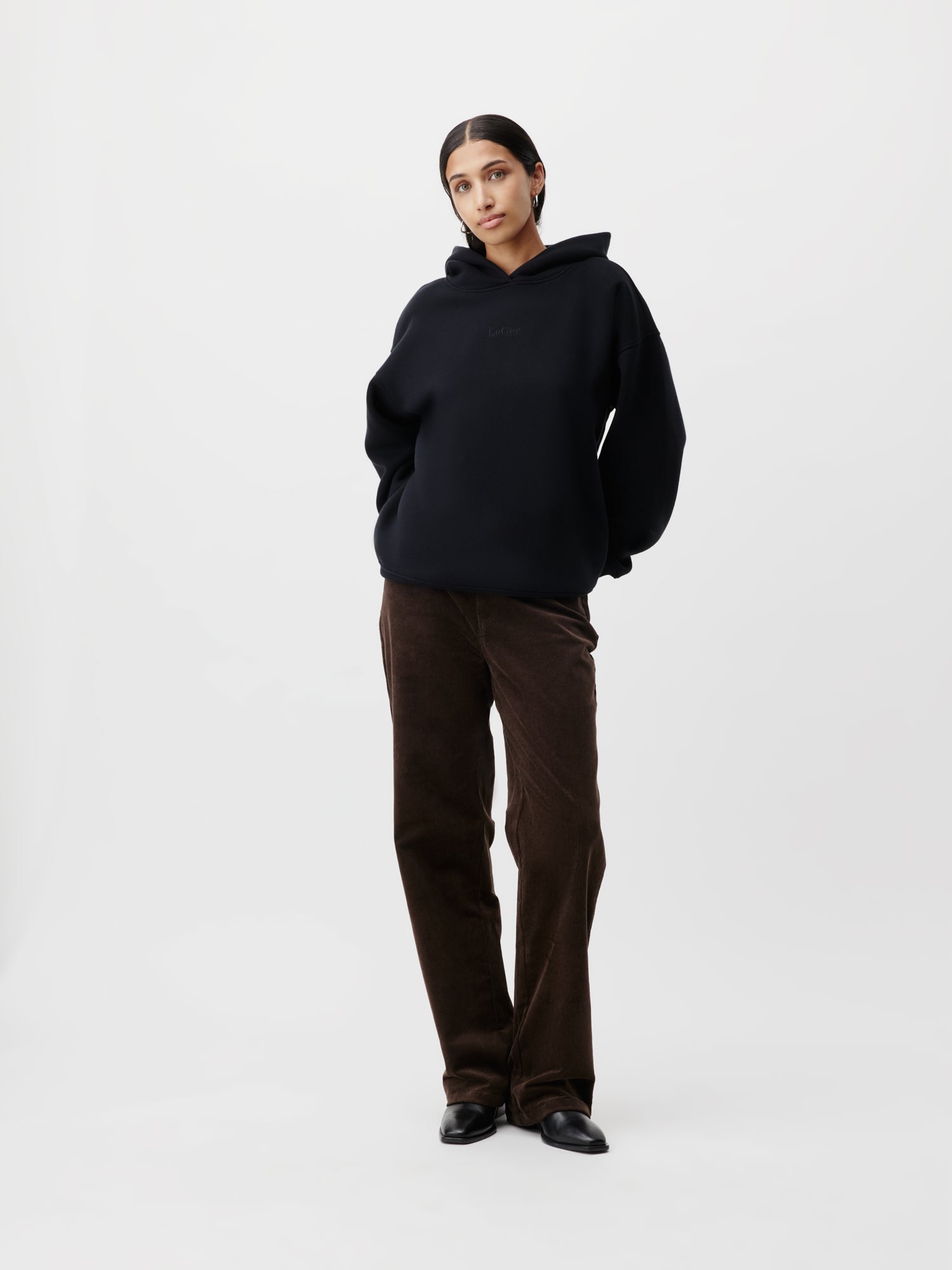 Eine Person im Abby Hoodie von LeGer by Lena Gercke und braunen Hosen steht selbstbewusst vor einem schlichten weißen Hintergrund. Die Hände stecken lässig in den Taschen, was die normale Passform ihrer Kleidung in Größe 36 unterstreicht, und sie trägt schwarze Schuhe.