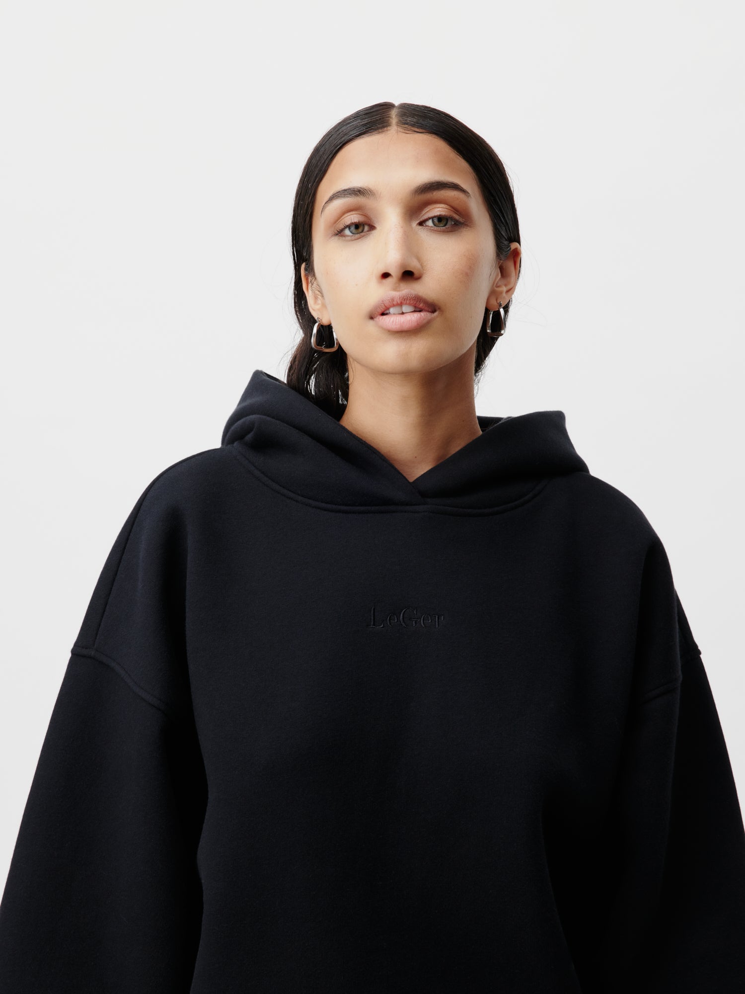 Eine Person mit zurückgebundenem, langem, dunklem Haar trägt einen schwarzen Abby Hoodie von LeGer by Lena Gercke, auf dem das Wort „Le Cop“ gestickt ist. Der Hoodie hat Größe 36 und lange Ärmel. Sie trägt Creolen und steht vor einem schlichten weißen Hintergrund.