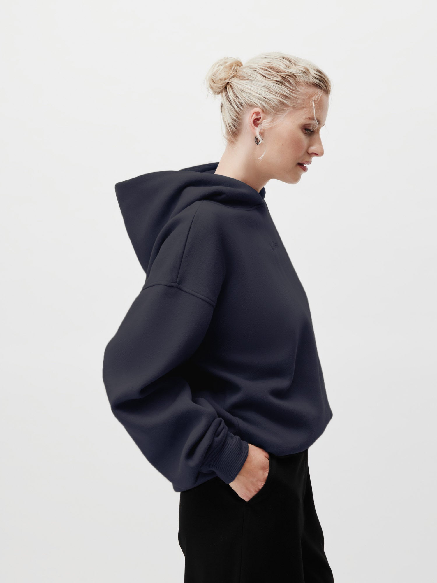 Eine blondhaarige Person mit Dutt trägt den Abby Hoodie von LeGer by Lena Gercke in dark navy (Größe 36) und eine schwarze Hose (normale Länge). Sie steht im Profil mit einer Hand in der Tasche vor einem schlichten weißen Hintergrund.
