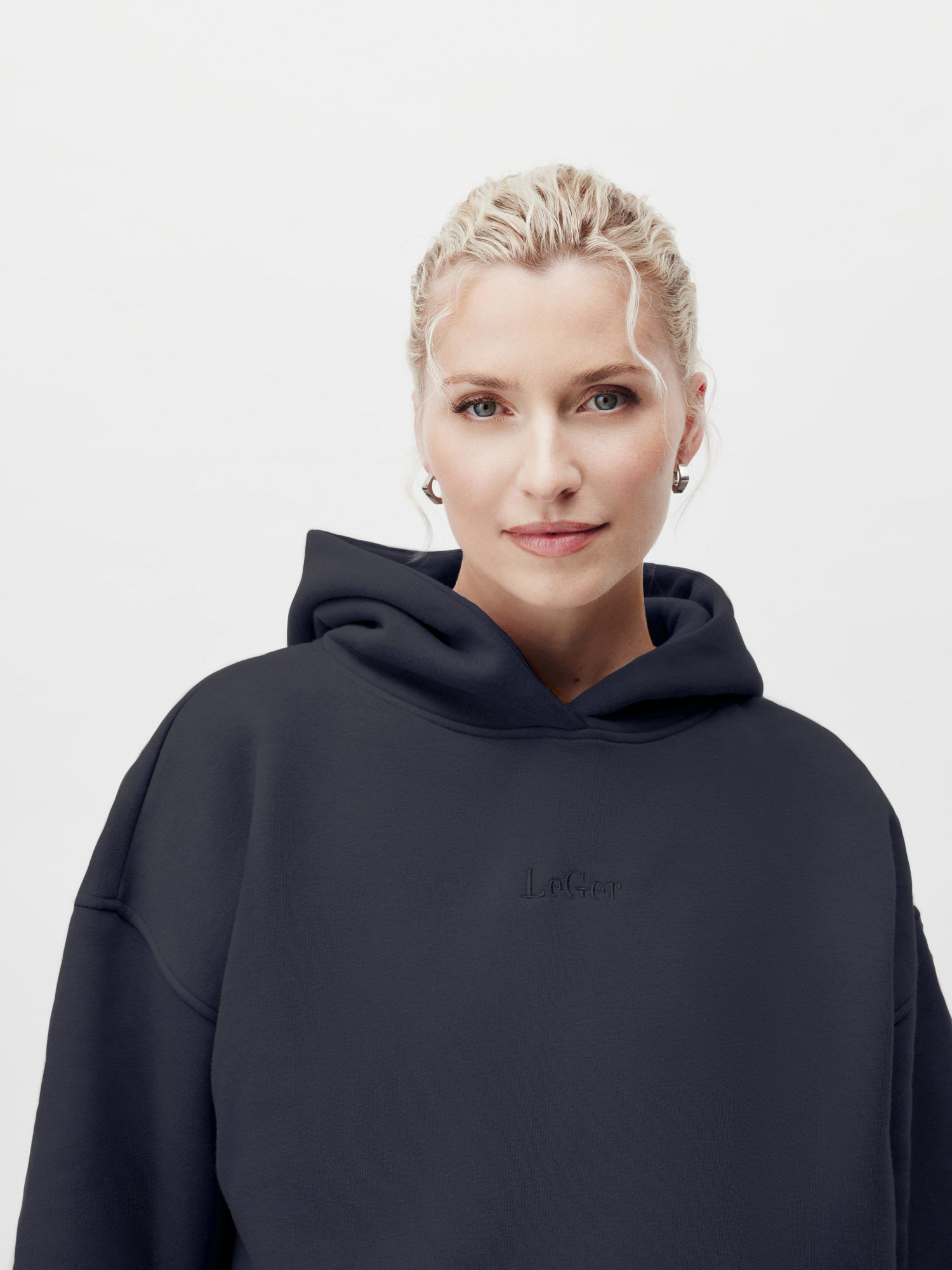 Eine Person mit kurzen, blonden, gewellten Haaren trägt den Abby Hoodie von LeGer by Lena Gercke in Größe 36 und kleine Ohrringe und blickt selbstbewusst mit einem leichten Lächeln in die Kamera vor einem schlichten, hellen Hintergrund.