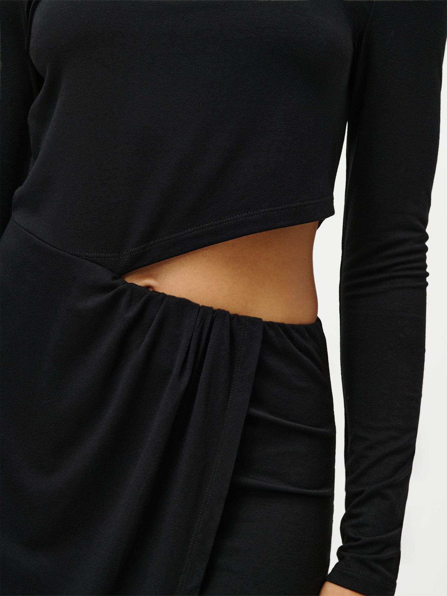 Eine Nahaufnahme zeigt eine Person, die das Kleid „Farina“ von LeGer by Lena Gercke trägt – ein schwarzes Minikleid mit einem asymmetrischen Ausschnitt an der Taille und einer diagonalen Drapierung über der Hüfte, die dezent Haut enthüllt.