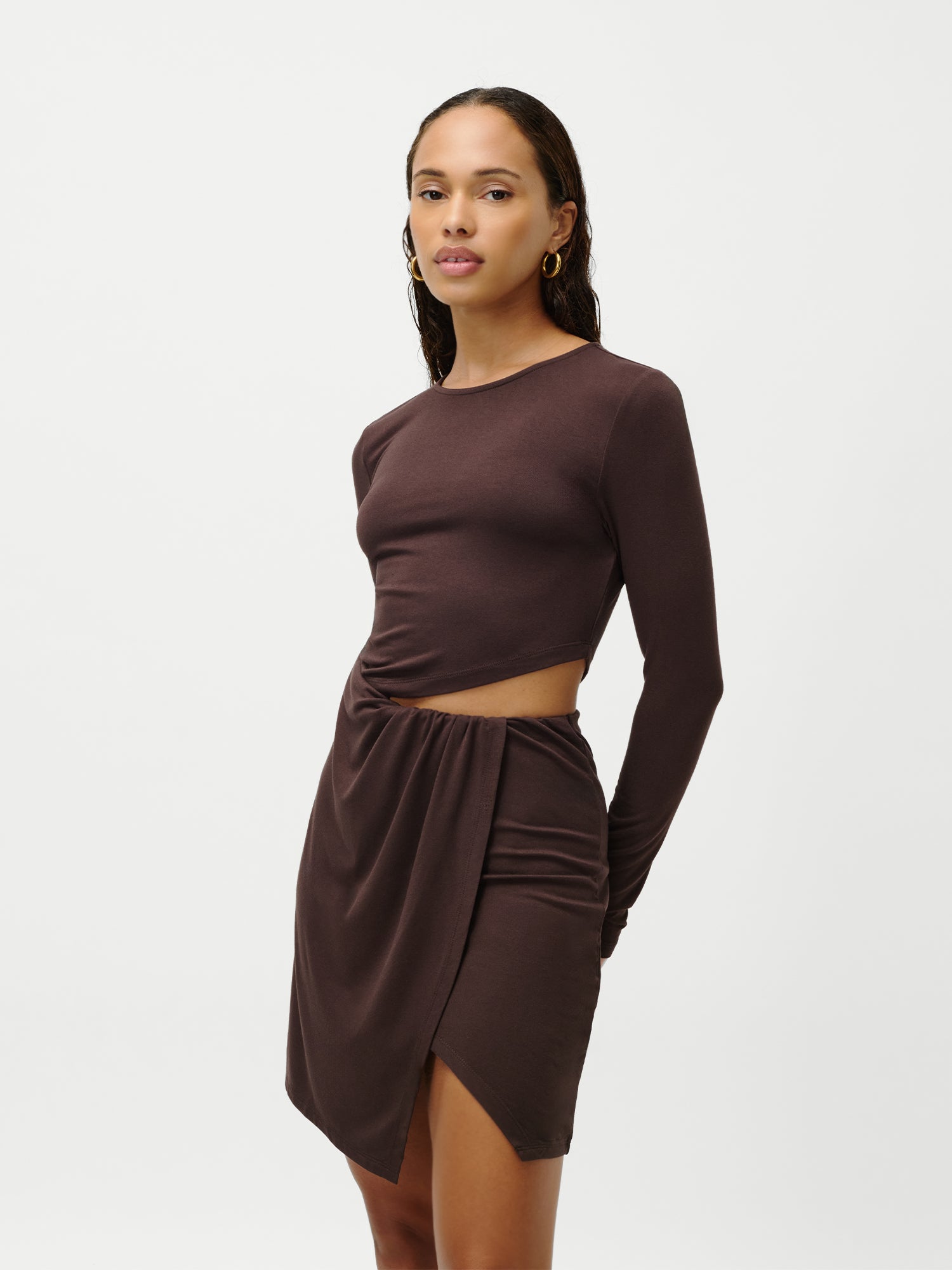 Eine Frau mit langen, dunklen Haaren trägt das Kleid „Farina“ von LeGer by Lena Gercke – ein figurbetontes, langärmeliges Minikleid mit einem seitlichen Ausschnitt an der Taille und einem asymmetrisch drapierten Rock – und steht dabei vor einem schlichten weißen Hintergrund.