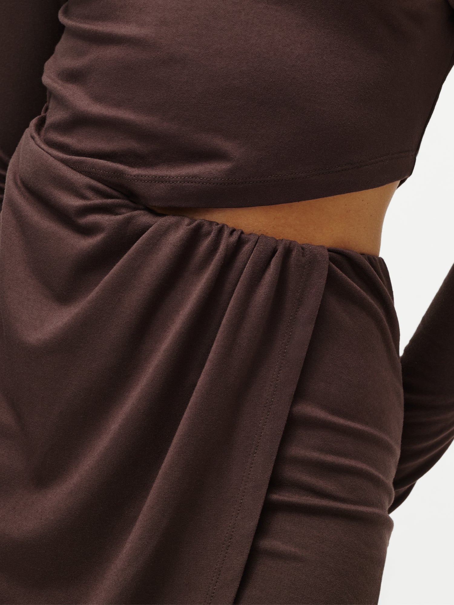 Eine Nahaufnahme einer Person, die das Kleid „Farina“ von LeGer by Lena Gercke trägt – ein dunkelbraunes, langärmeliges Minikleid mit Cut-outs, gerafftem Stoff an der Taille und einem dezenten Blick auf den Bauch.