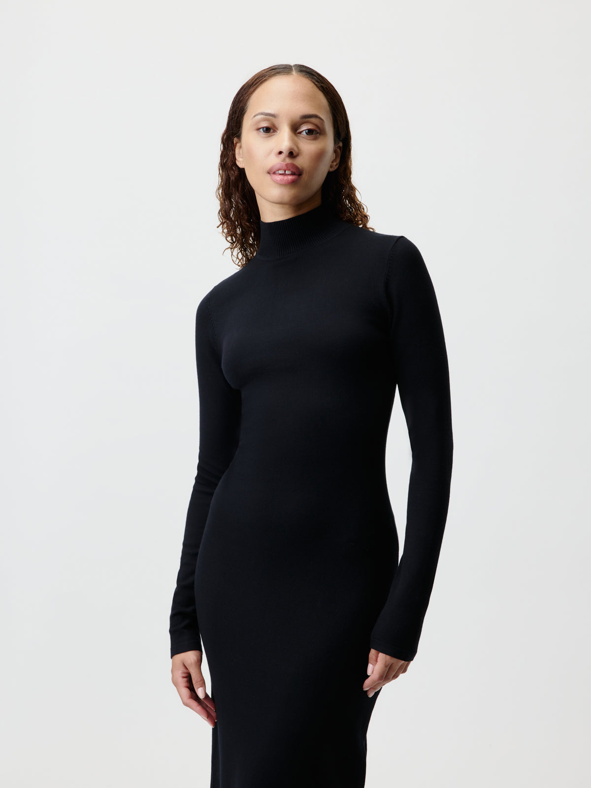 Eine Person mit schulterlangen Locken trägt das „Imen“-Kleid von LeGer by Lena Gercke mit langen Ärmeln und schwarzem Rollkragen und steht vor einem schlichten, hellen Hintergrund, wobei sie mit neutralem Gesichtsausdruck in die Kamera blickt.