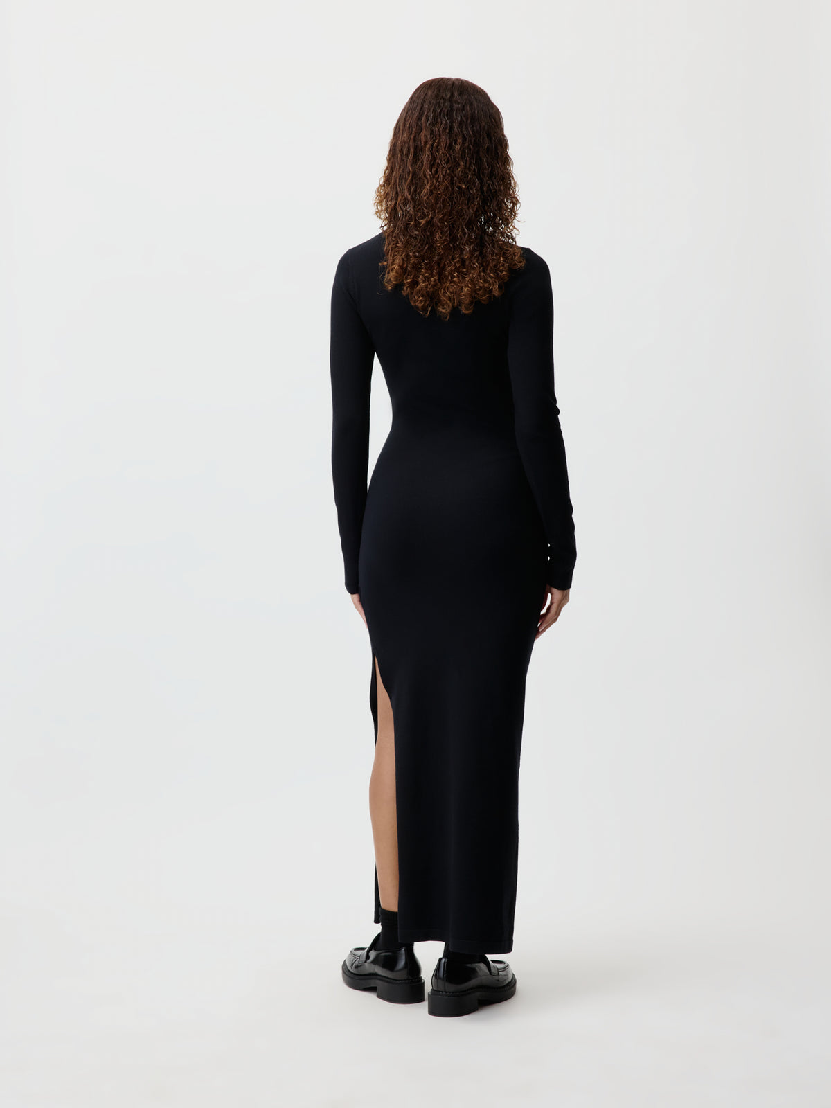 Eine Person mit lockigem Haar, die das langärmelige, knöchellange schwarze „Imen“-Kleid von LeGer by Lena Gercke mit seitlichem Schlitz trägt, steht mit dem Rücken zur Kamera vor einem schlichten weißen Hintergrund.
