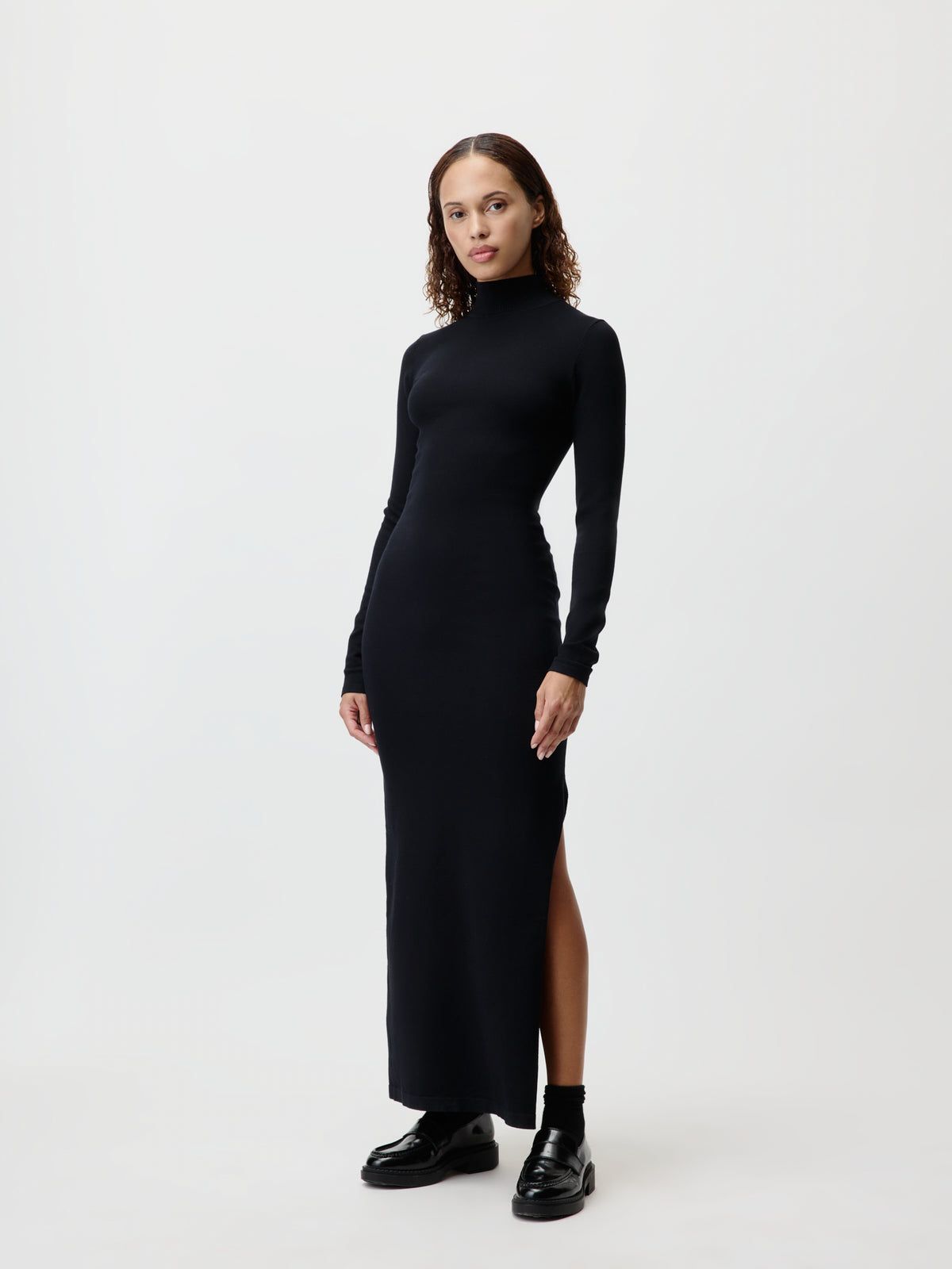 Eine Frau mit lockigem Haar trägt ein langärmeliges, schwarzes „Imen“-Maxikleid mit Rollkragen von LeGer by Lena Gercke, das einen Seitenschlitz aufweist, dazu schwarze Schuhe, und steht vor einem schlichten weißen Hintergrund.