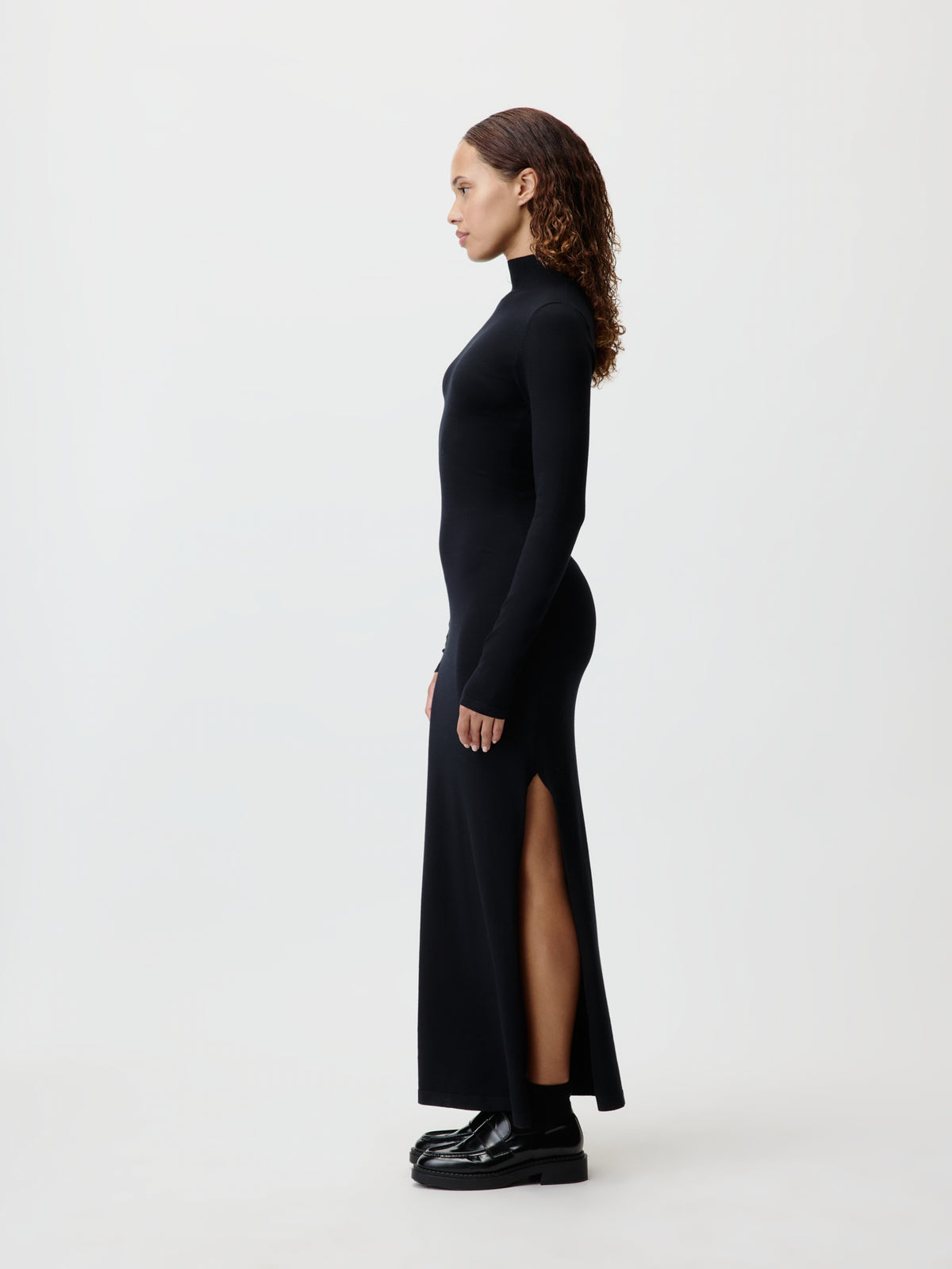 Eine Frau steht im Profil vor einem schlichten weißen Hintergrund und trägt das langärmelige, knöchellange schwarze „Imen“-Kleid von LeGer by Lena Gercke mit seitlichem Schlitz, dazu schwarze Schuhe.