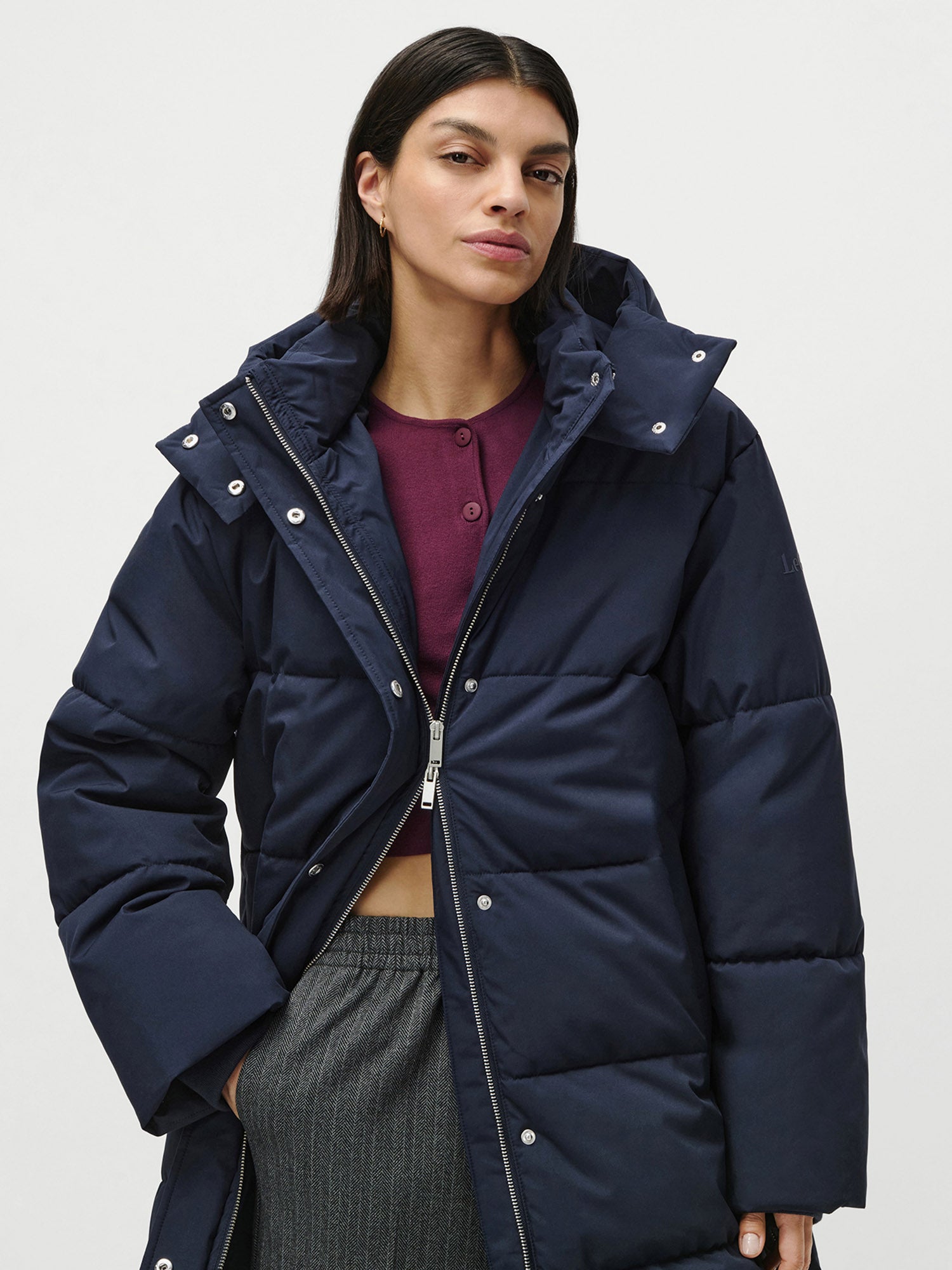 Eine Frau mit langen dunklen Haaren trägt den Bessy Coat von LeGer by Lena Gercke, eine marineblaue Pufferjacke mit langen Ärmeln und normaler Länge, über einem weinroten Oberteil und einer grauen Hose, vor einem schlichten hellen Hintergrund.
