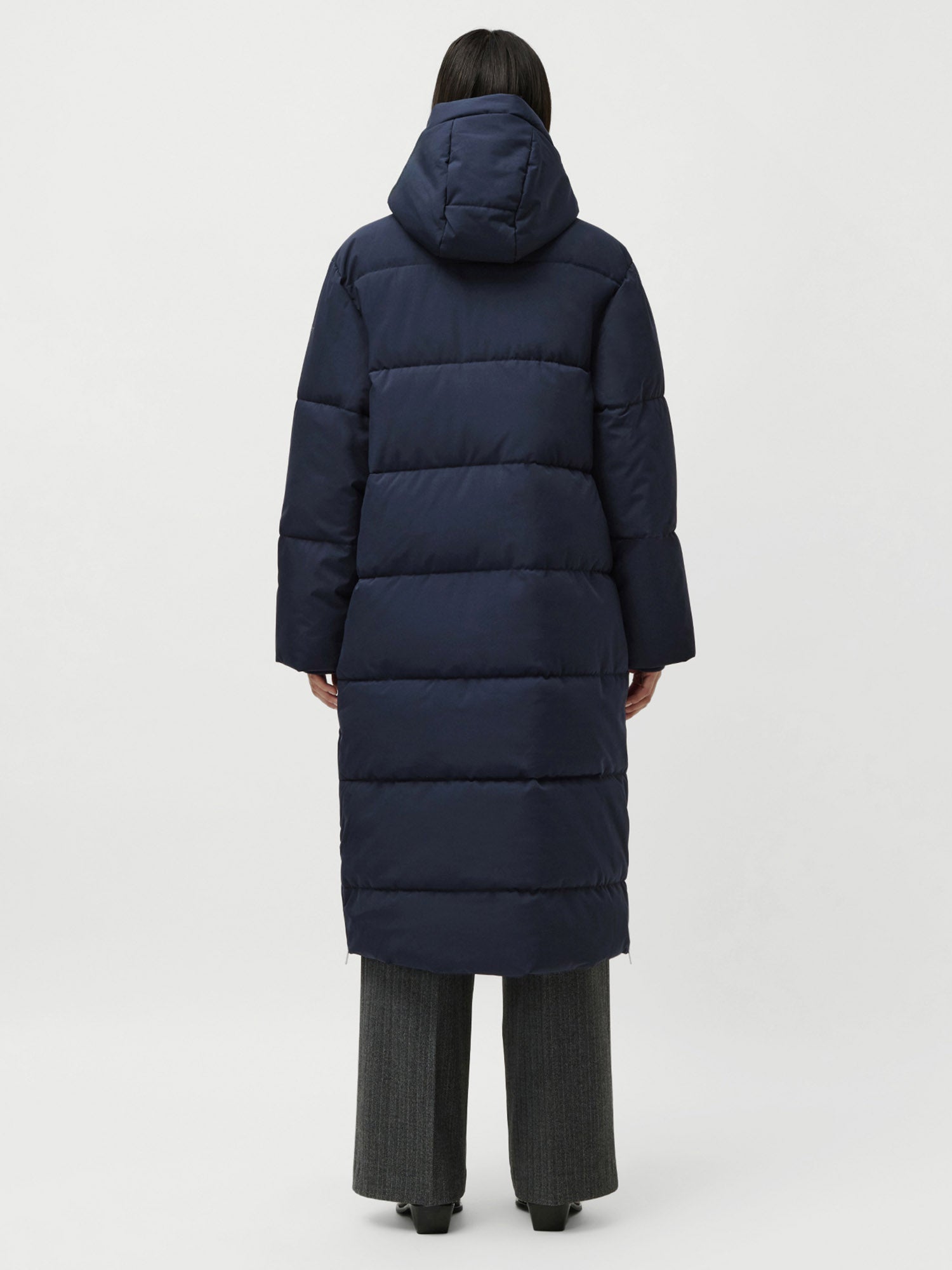 Eine Person mit dunklen Haaren ist von hinten zu sehen, die den LeGer by Lena Gercke Bessy Coat trägt, einen marineblauen Puffmantel mit Kapuze in normaler Länge und lockerer Passform, kombiniert mit einer grauen Hose mit weitem Bein und schwarzen Schuhen vor einem schlichten weißen Hintergrund.