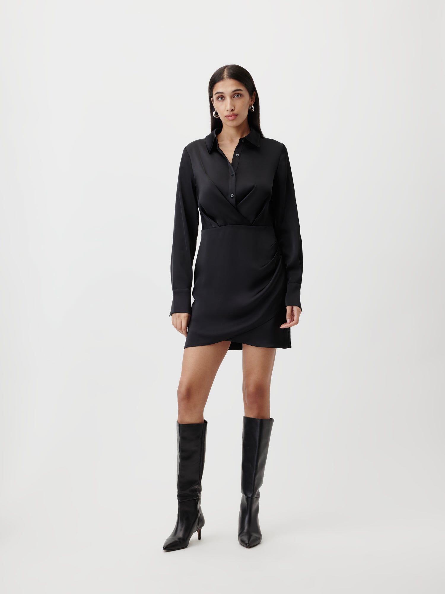 Eine Frau mit langen dunklen Haaren steht vor einem weißen Hintergrund, trägt das LeGer by Lena Gercke Dress Model "Braelyn" von LeGer by Lena Gercke und kniehohe schwarze Stiefel und schaut mit neutralem Blick in die Kamera.