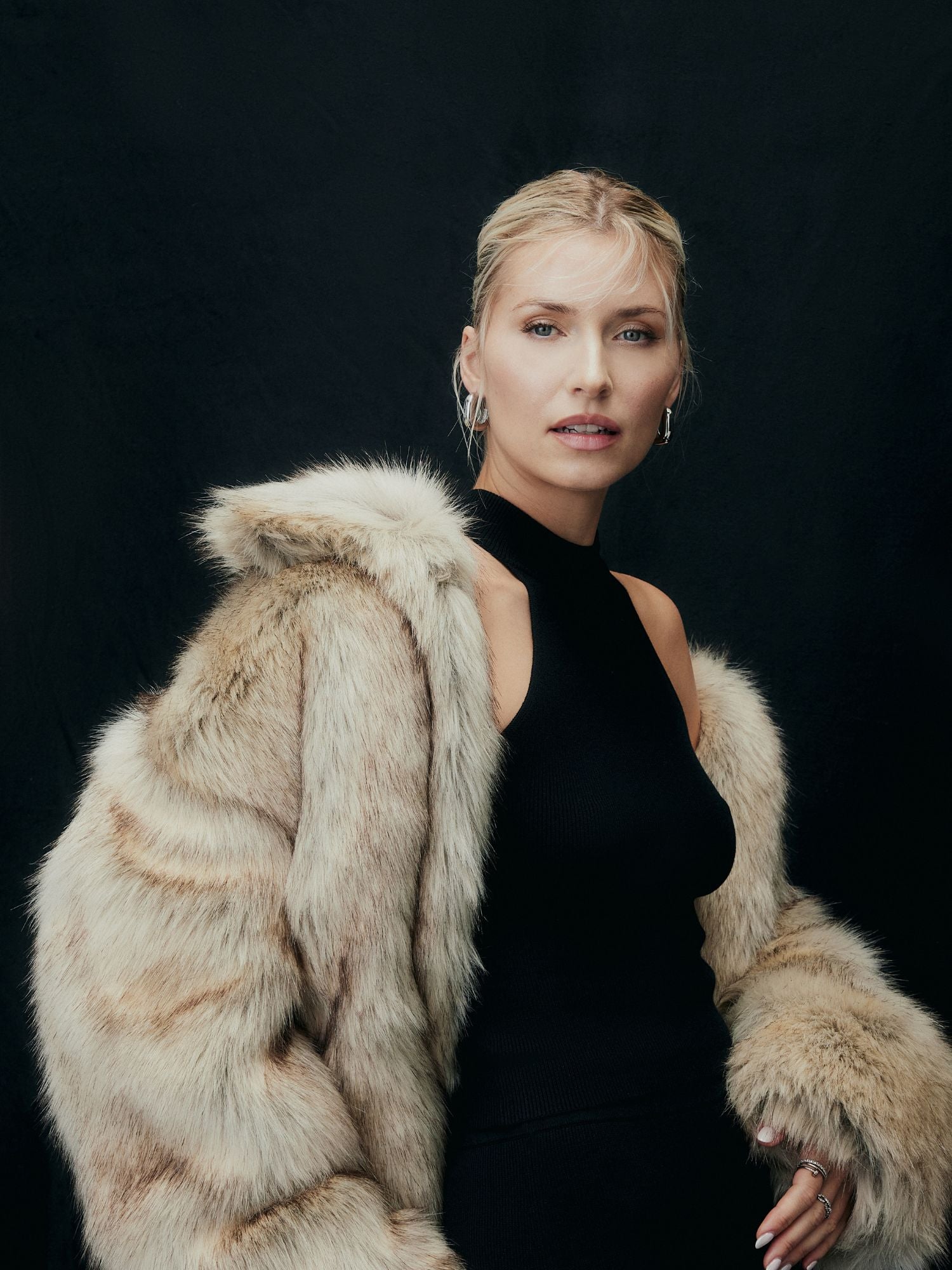 Eine Frau mit blondem Haar in einer lockeren Hochsteckfrisur trägt ein schwarzes ärmelloses Oberteil und drapiert die LeGer by Lena Gercke Fergie Jacket über ihre Schultern. Sie posiert vor einem dunklen Hintergrund.