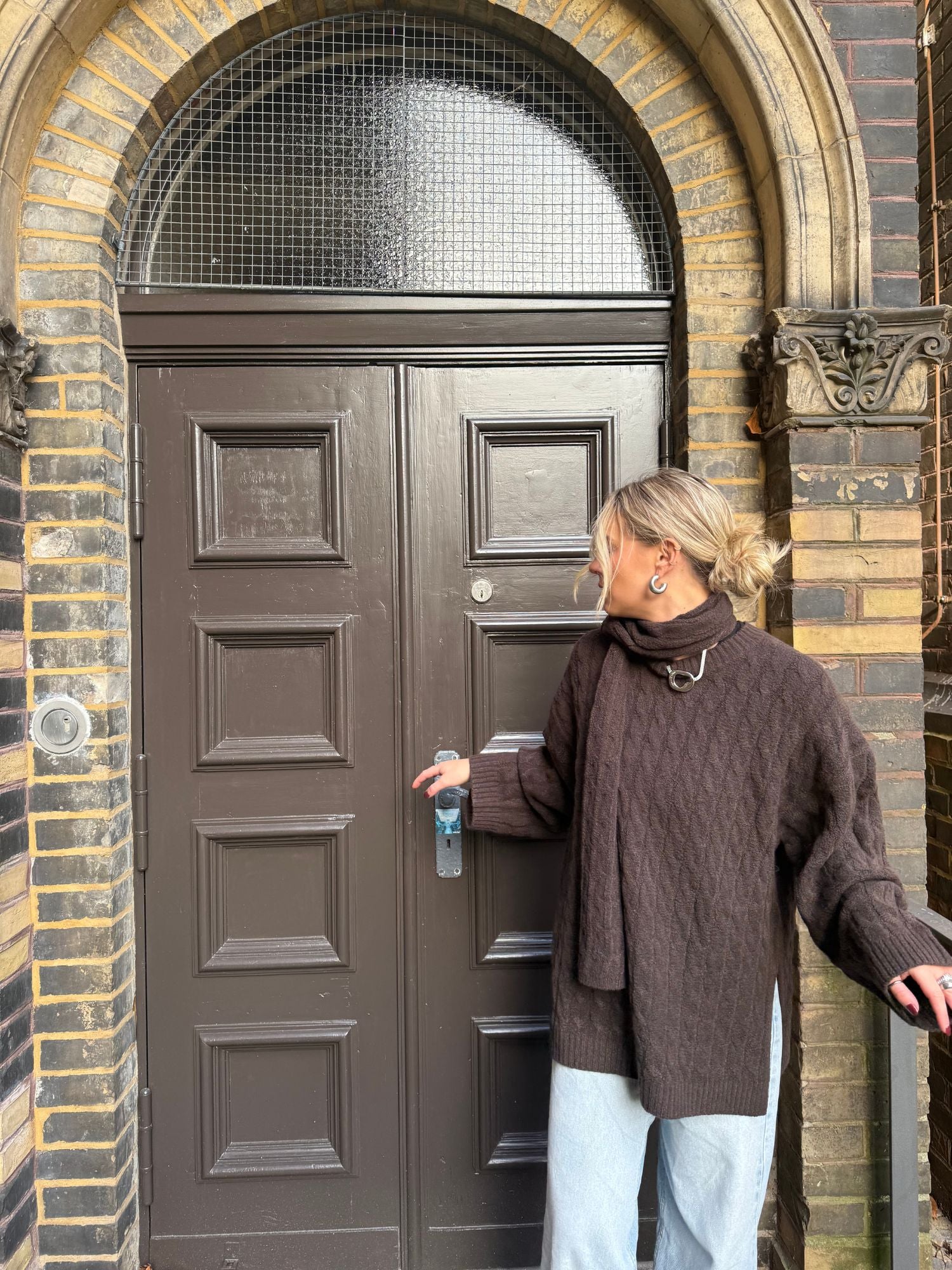Eine Person trägt den dunkelbraunen Jamila Jumper von LeGer by Lena Gercke in Größe 36 und helle Jeans, steht vor einer braunen, getäfelten Tür im Rundbogen, blickt zur Seite und hält den Türgriff.