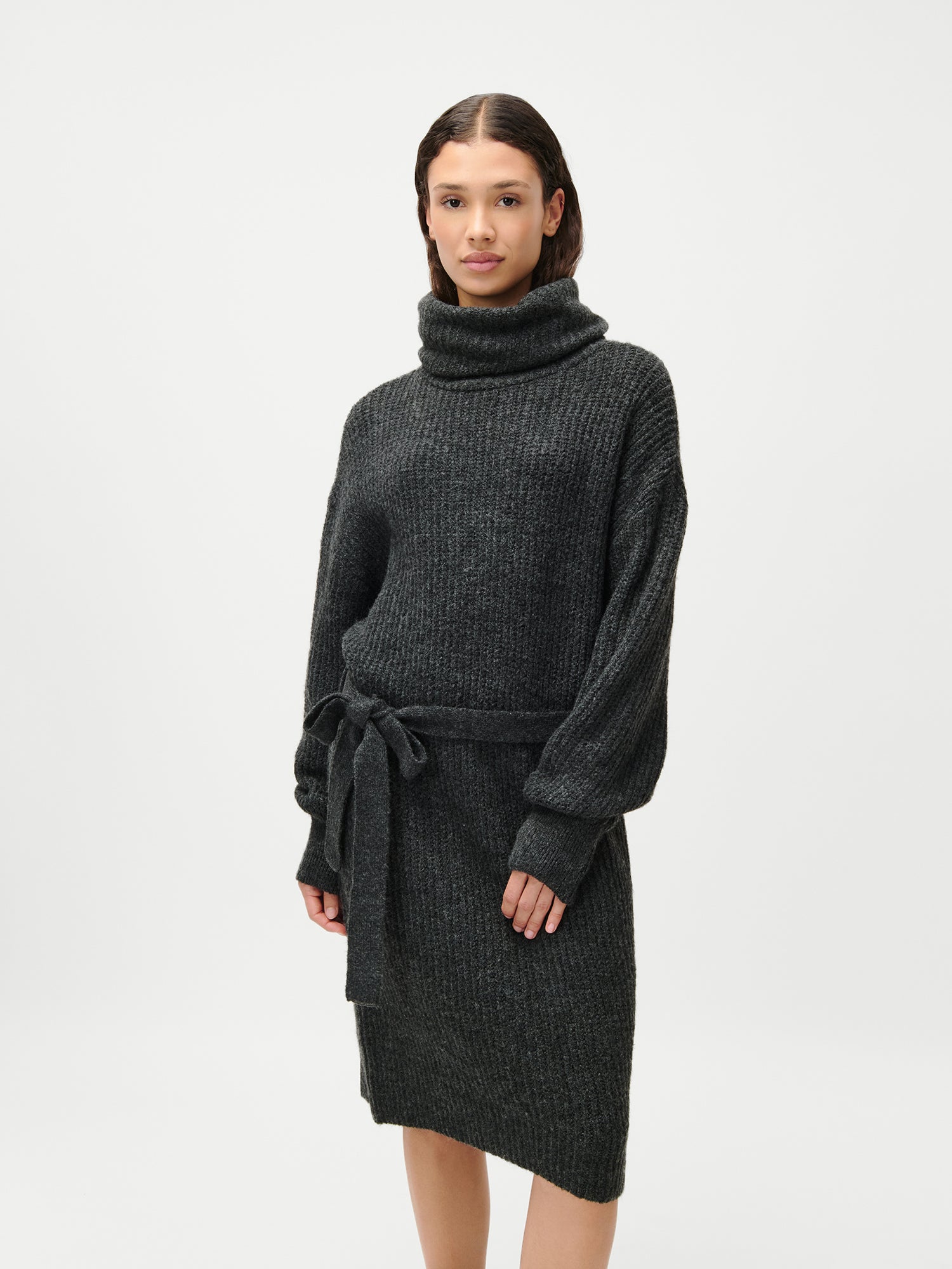 Eine Frau trägt das Colleen Dress von LeGer by Lena Gercke: ein knielanges, dunkelgraues, geripptes Langarmkleid mit Rollkragen und Taillengürtel in Größe 36 vor schlicht weißem Hintergrund.