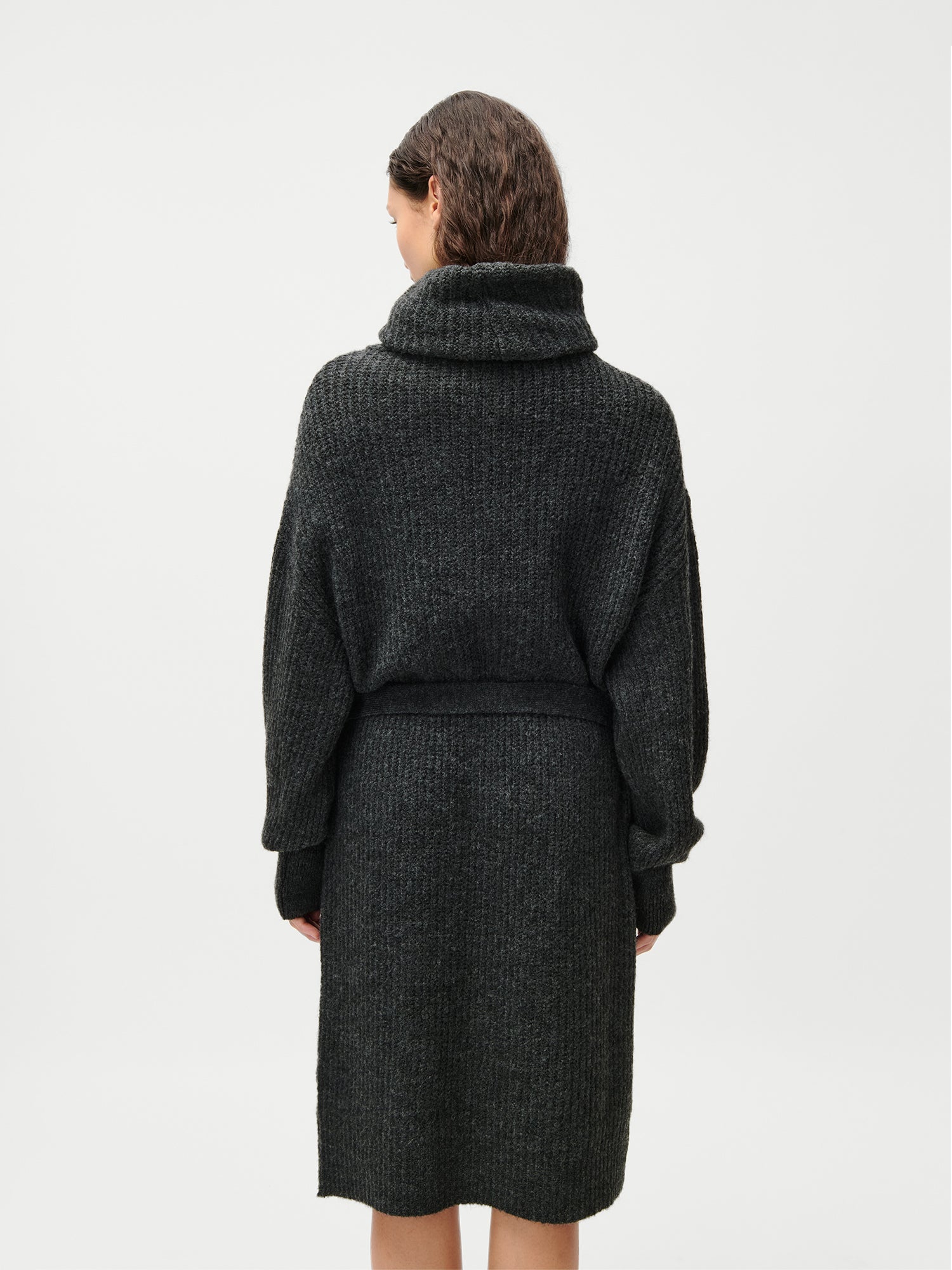 Eine Frau mit gewelltem braunem Haar blickt nach vorne und trägt das Colleen Dress von LeGer by Lena Gercke - ein knielanges, dunkelgraues, langärmeliges Kleid in Größe 36 mit Rollkragen und Taillengürtel - vor einem schlichten weißen Hintergrund.