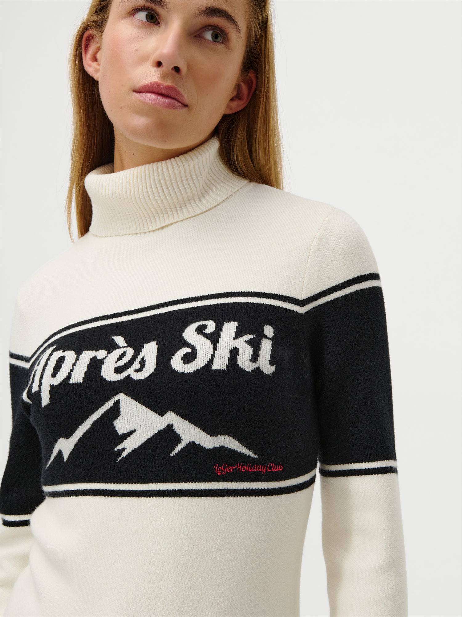 Eine Frau trägt den Floriane Jumper von LeGer by Lena Gercke, einen cremefarbenen, langärmeligen Rollkragenpullover in Größe 36 mit einem schwarzen Brustband, auf dem "Après Ski" in Weiß und eine Berggrafik zu sehen sind, sowie "Aspen Holiday Club" in Rot darunter gestickt.