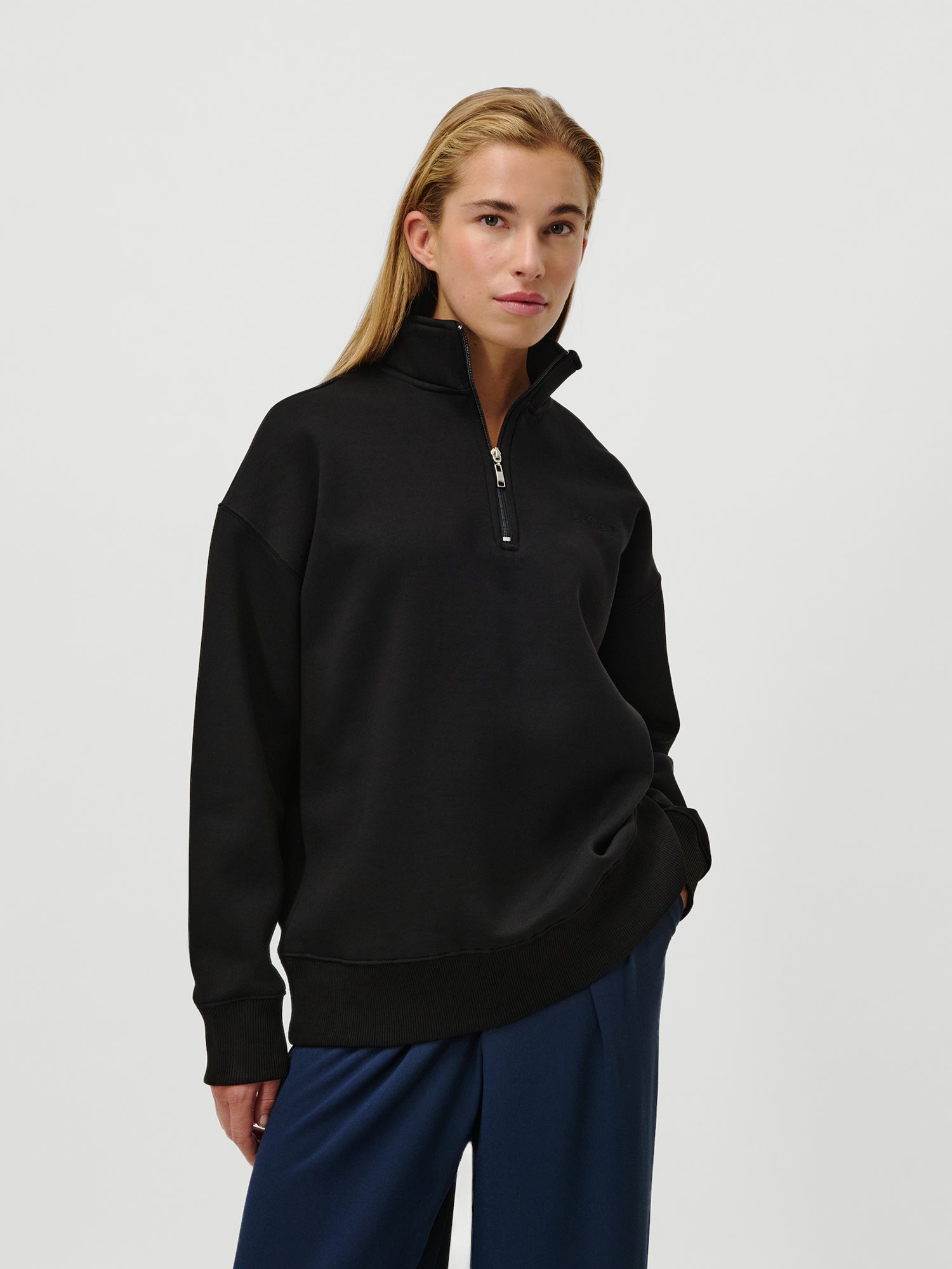 Eine Person mit langen blonden Haaren trägt den LeGer by Lena Gercke Jenny Sweater, einen schwarzen Viertel-Reißverschluss-Pullover mit langen Ärmeln und normaler Länge, gepaart mit einer dunkelblauen Hose, und steht selbstbewusst vor einem weißen Hintergrund, eine Hand in der Tasche.