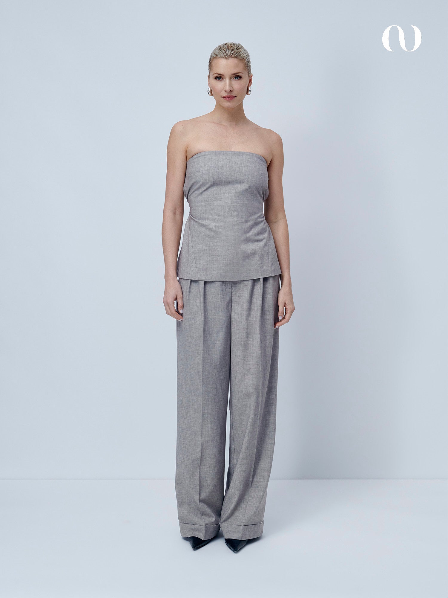 Eine Frau mit blondem Haar posiert vor einem hellen Hintergrund im LeGer by Lena Gercke Jale Top, einem trägerlosen grauen Modell, das mit einer passenden Hose mit weitem Bein und verstellbarem Bund kombiniert wird. Schwarze spitze Schuhe und ein weißes abstraktes Logo vervollständigen den Look.