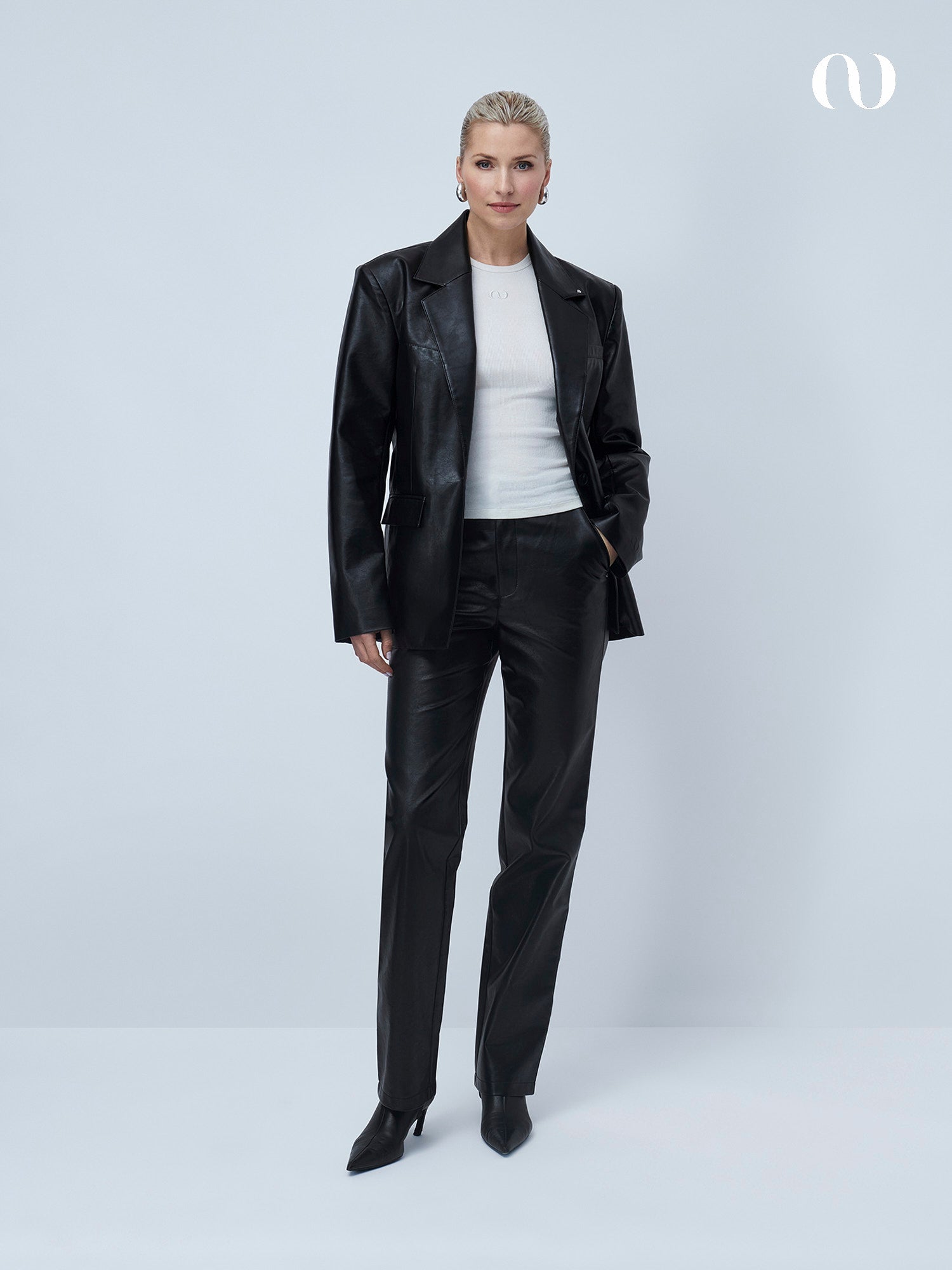 Eine Frau mit zurückgegelten blonden Haaren und Ohrringen posiert selbstbewusst in der Insa Pants von LeGer by Lena Gercke, gestylt mit einem schwarzen Lederblazer, einem weißen Top und schwarzen Schuhen mit Absatz vor einem schlichten, hellen Hintergrund.