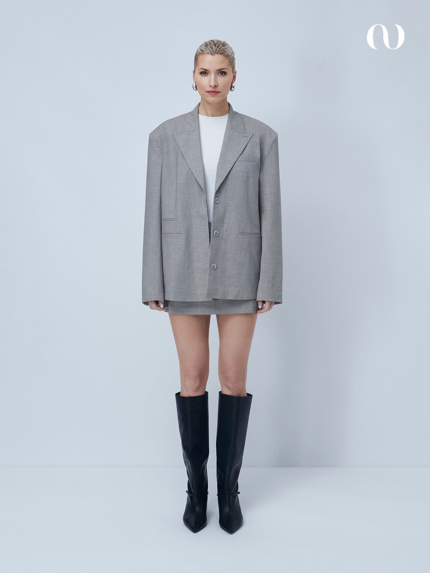 Eine Frau mit kurzen, zurückgegelten blonden Haaren und einem neutralen Gesichtsausdruck steht vor einem hellgrauen Hintergrund und trägt den Jale Blazer von LeGer by Lena Gercke, ein weißes Minikleid und kniehohe schwarze Stiefel.