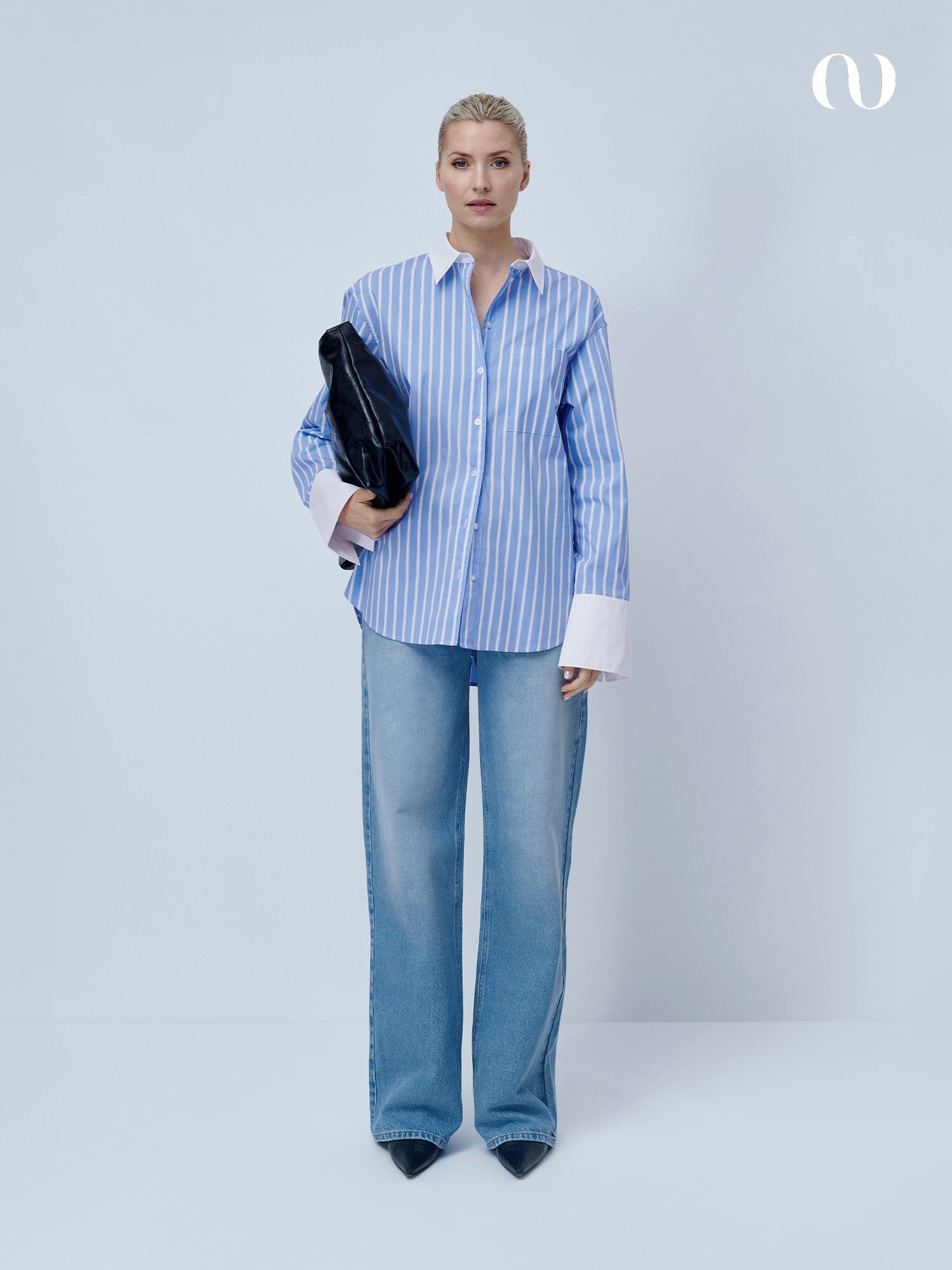 Eine Person steht vor einem schlichten hellen Hintergrund und trägt ein blau-weiß gestreiftes Hemd, schwarze spitze Schuhe, eine dunkle Tasche und eine hellblaue Rhonda-Hose mit weitem Bein und mittlerer Leibhöhe von LeGer by Lena Gercke.