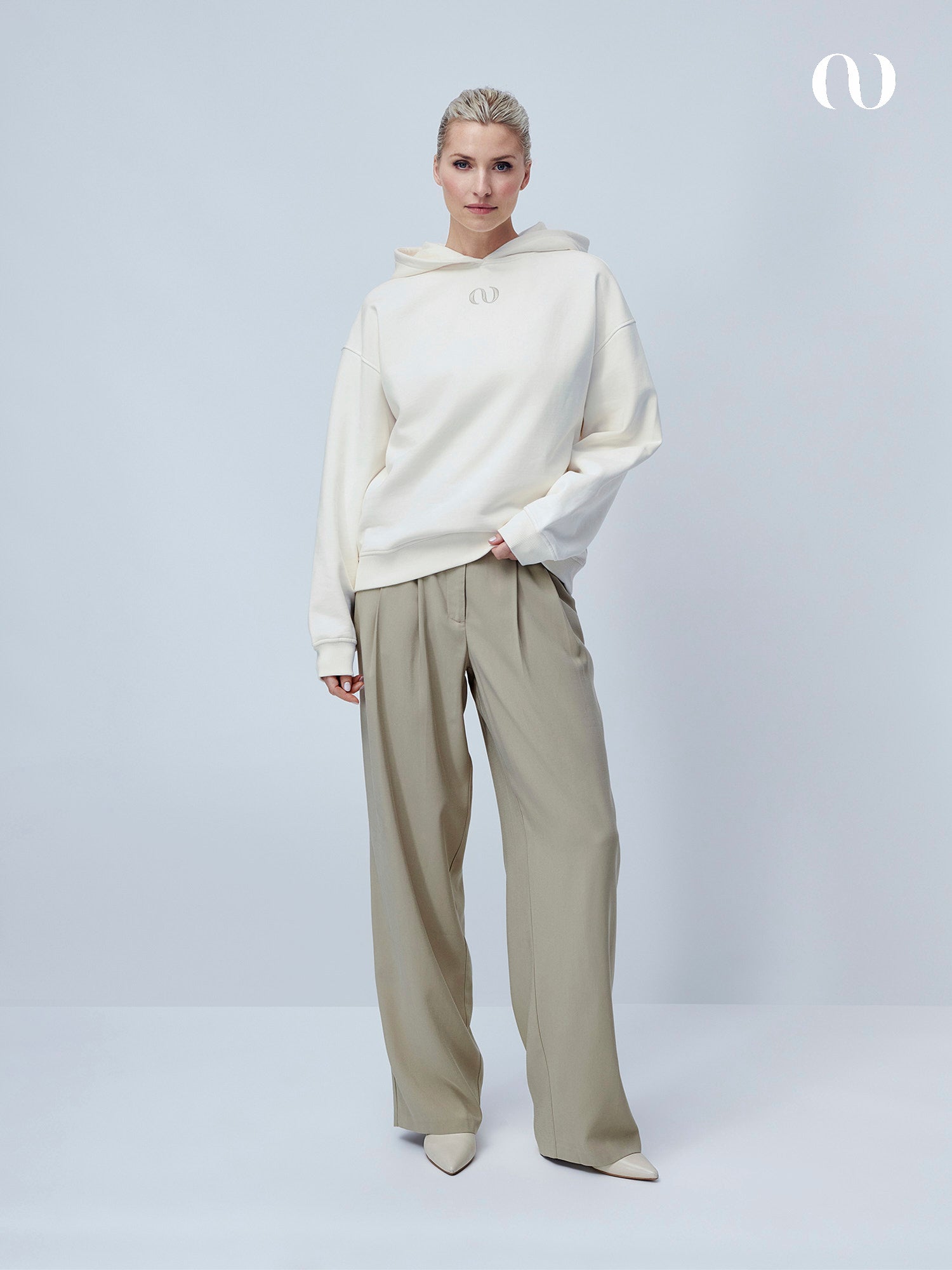 Eine Frau mit kurzen blonden Haaren und neutralem Gesichtsausdruck steht vor einem hellgrauen Hintergrund und trägt den übergroßen cremefarbenen Talea Hoodie von LeGer by Lena Gercke, eine weite beigefarbene Hose und spitze Schuhe. Ein minimalistisches Logo befindet sich in der oberen rechten Ecke.