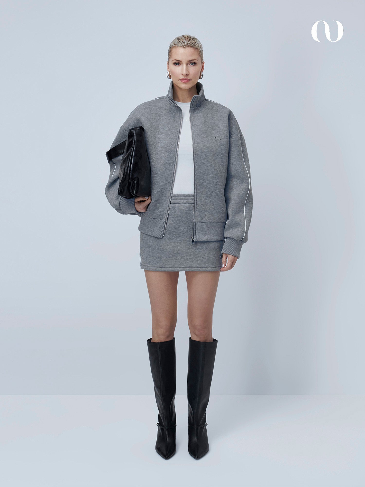 Eine Frau posiert vor einem hellen Hintergrund in dem LeGer by Lena Gercke Lilou Set - einer grauen Sweatjacke und einem passenden Minirock - mit einem weißen Top, schwarzen kniehohen Stiefeln und einer schwarzen Clutch unter dem Arm.