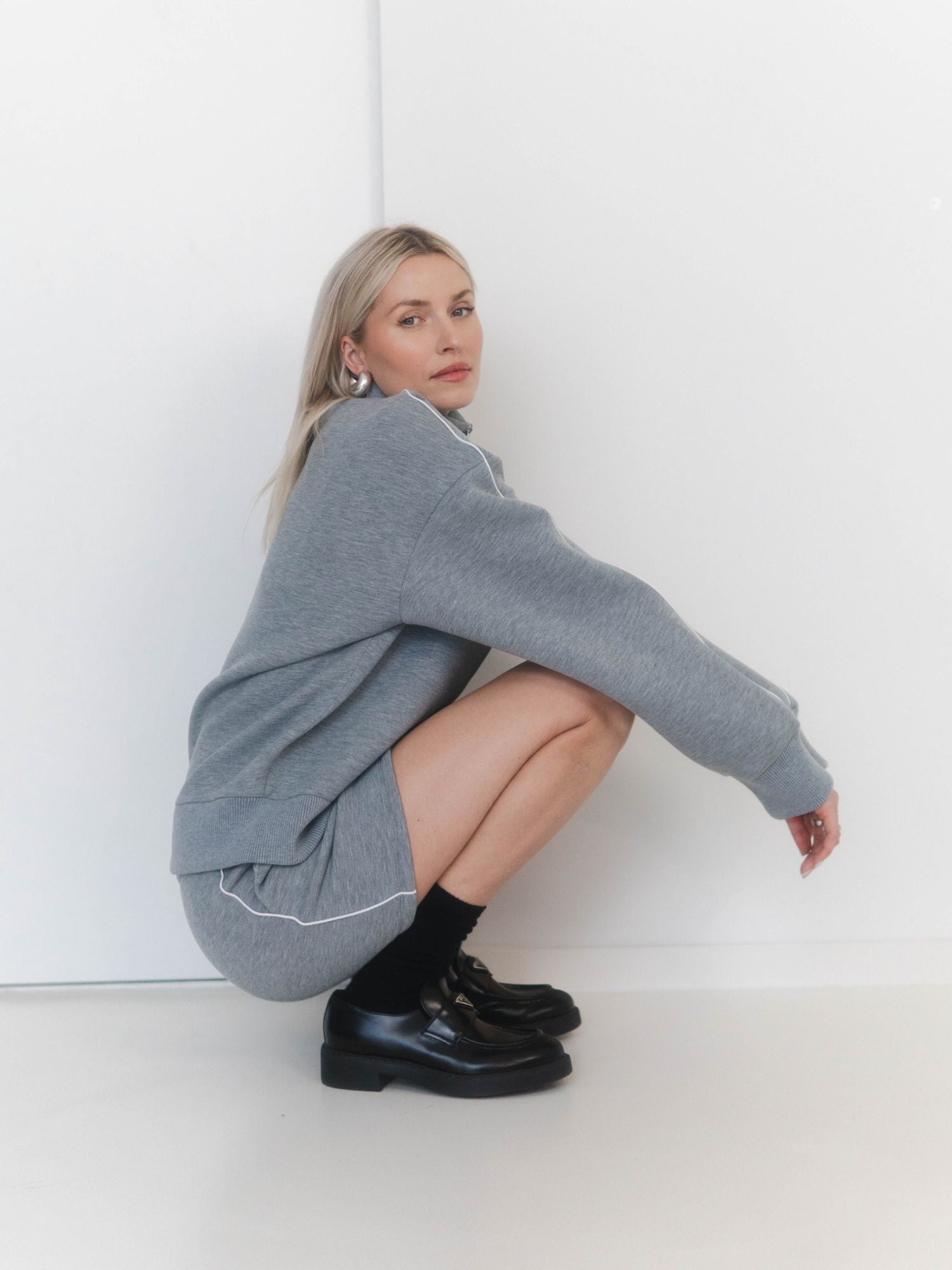 Eine Frau mit blonden Haaren hockt vor einer weißen Wand, trägt das graue Lilou-Set von LeGer by Lena Gercke, gepaart mit schwarzen Socken und Schuhen, und schaut ruhig in die Kamera.