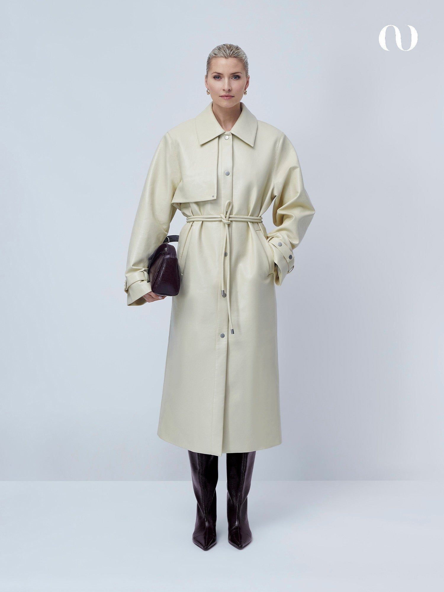 Eine Frau mit blondem, zurückgestyltem Haar und neutralem Gesichtsausdruck steht vor einem unifarbenen Hintergrund und trägt den Birka Coat von LeGer by Lena Gercke, gepaart mit dunklen kniehohen Stiefeln und einer dunklen Handtasche.