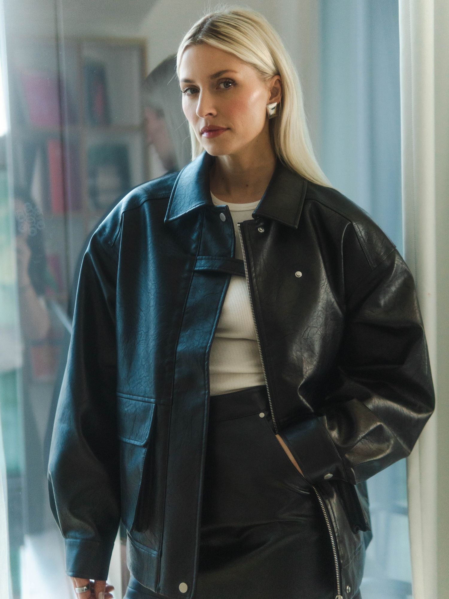 Eine Frau mit langen blonden Haaren trägt die LeGer by Lena Gercke Davinia Jacket und einen Rock über einem weißen Oberteil. Sie steht in einem Haus und hat einen neutralen Gesichtsausdruck. Im Hintergrund sind Bücherregale und ein Fenster zu sehen.