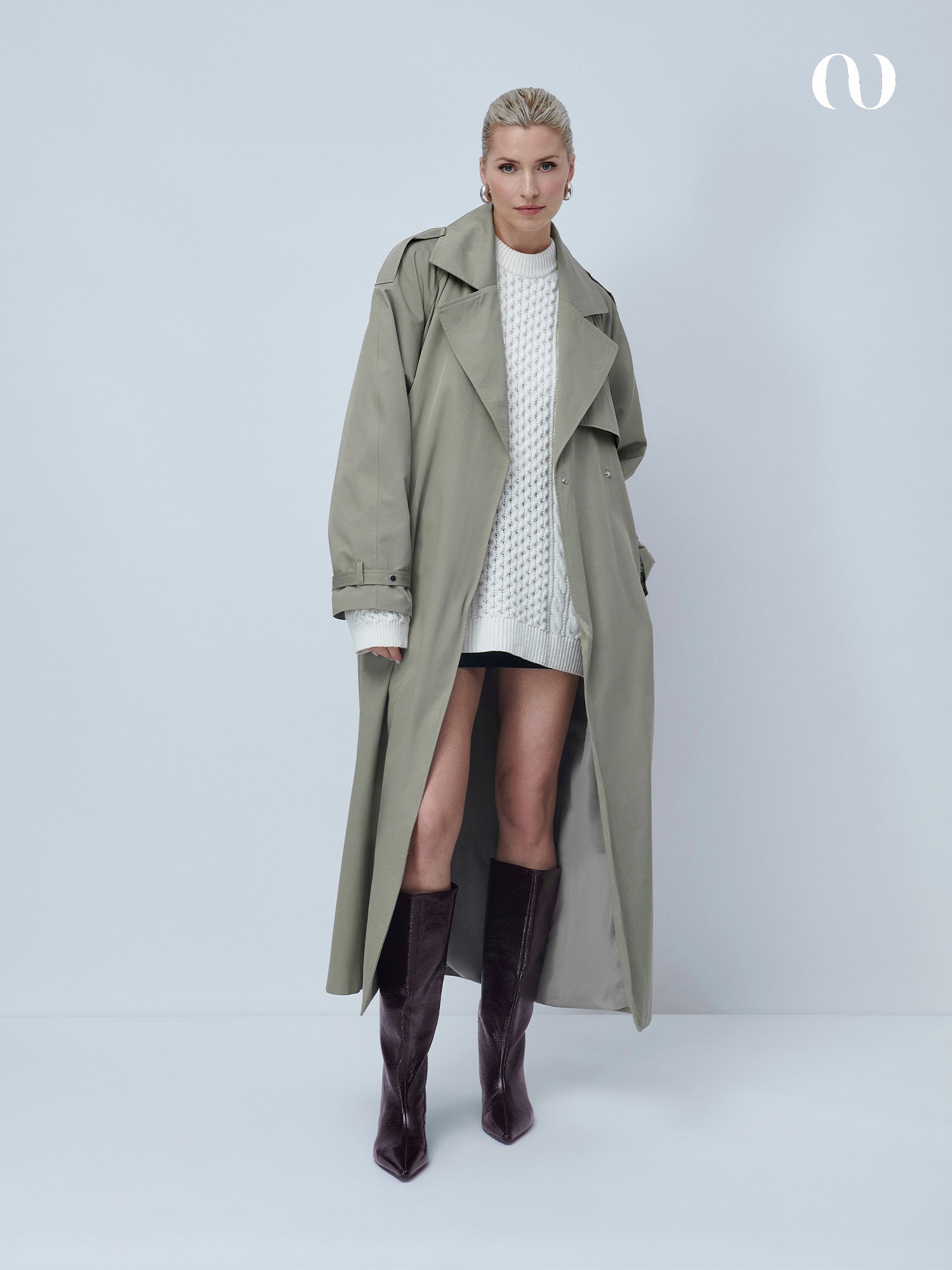 Eine Frau steht vor einem schlichten Hintergrund und trägt den übergroßen hellgrauen Elena Coat von LeGer by Lena Gercke über einem weißen strukturierten Pulloverkleid und hohen dunkelbraunen Samtstiefeln.