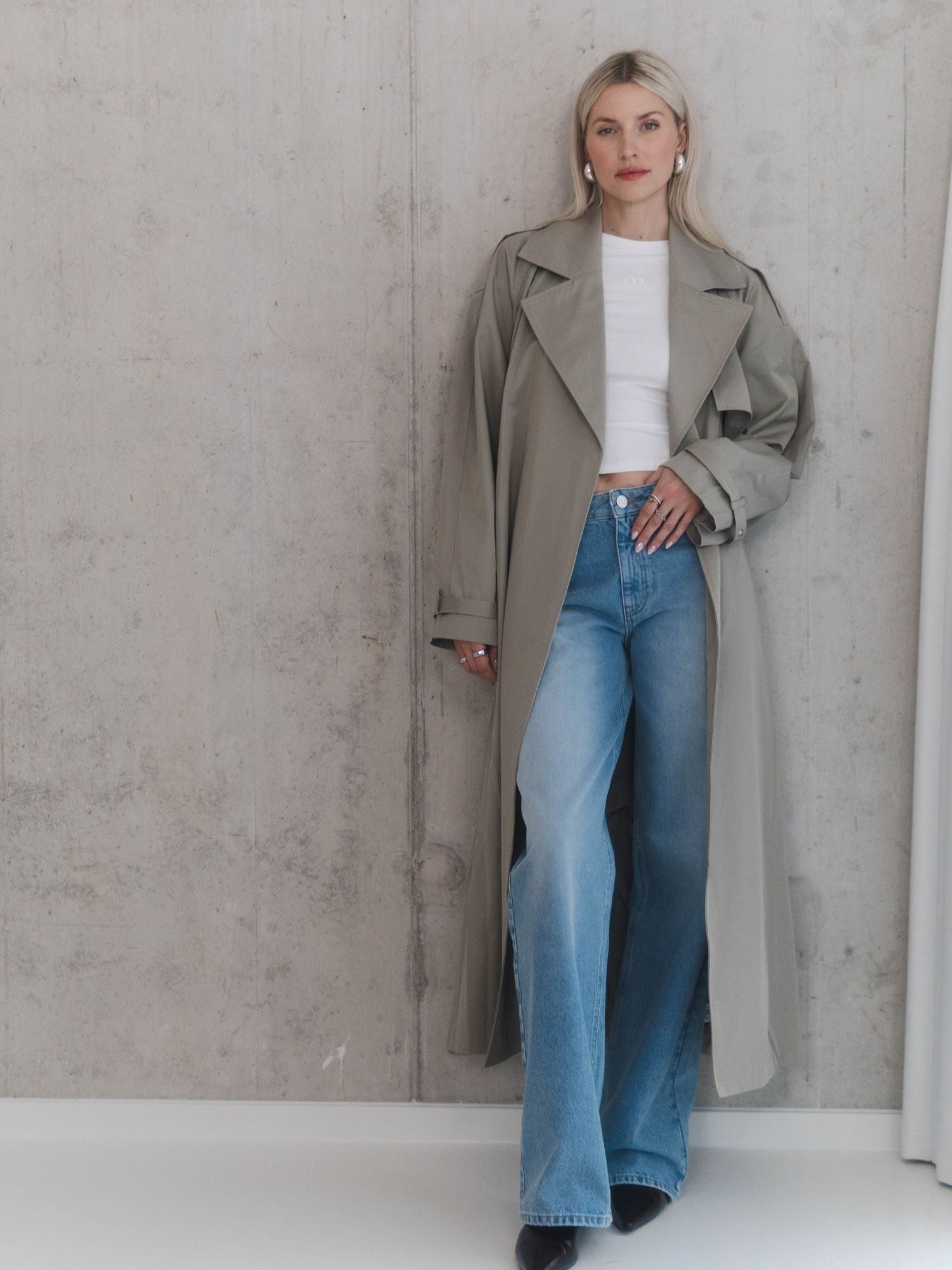 Eine Frau posiert selbstbewusst vor einer Betonmauer. Sie trägt den hellgrauen Mantel Elena von LeGer by Lena Gercke in einer lockeren Passform über einem weißen Crop-Top und blauen Schlagjeans. Sie hat langes blondes Haar und trägt dazu Perlenohrringe.